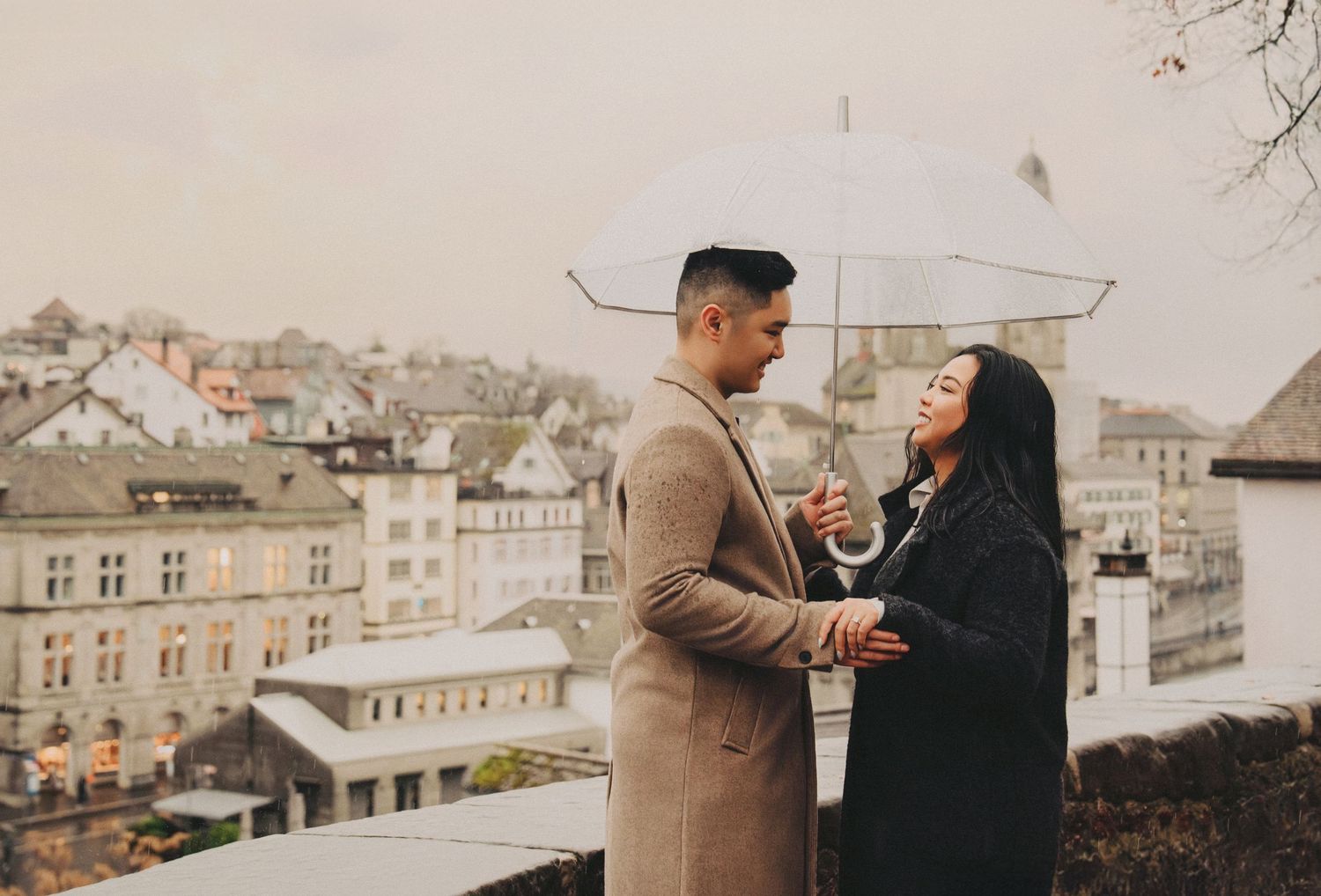Smiling couple sharing an umbrella on a rainy day with European city rooftops behind.