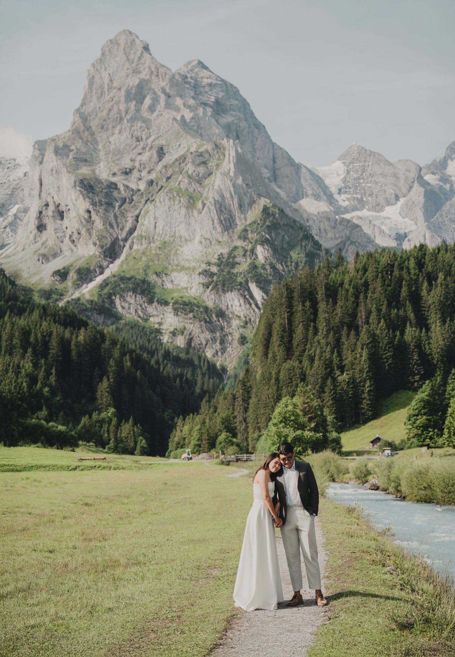Wedding couple embracing on a mountain path beside a turquoise river with towering rocky peaks in the background.