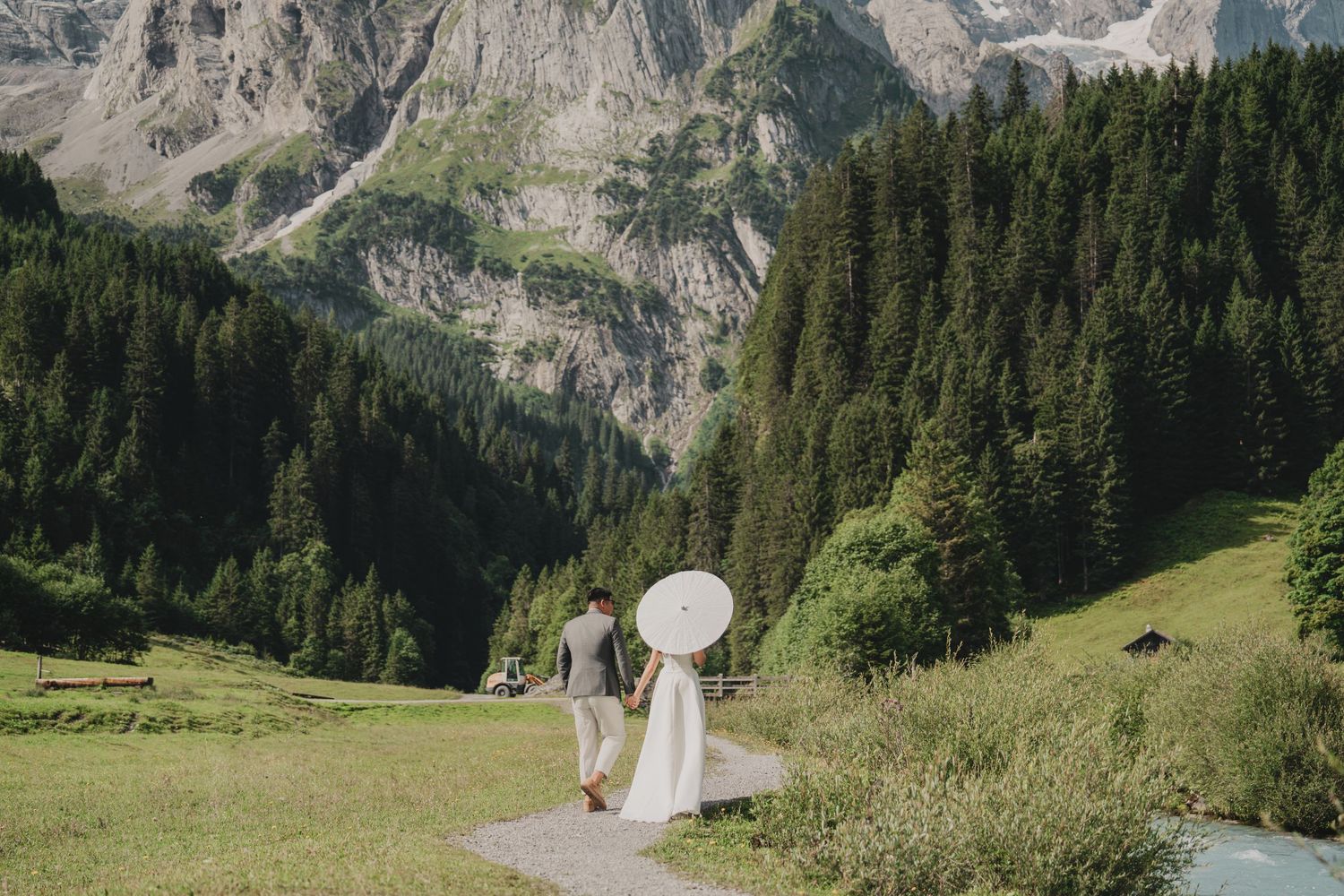 Newlywed couple strolling through a lush Alpine meadow with a white parasol and dramatic mountain scenery.