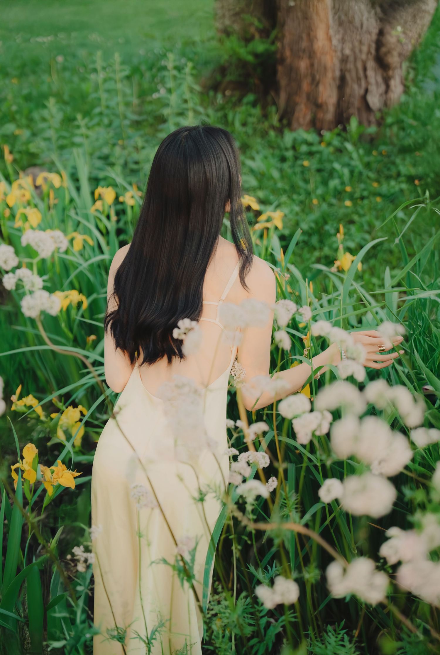 Woman in yellow dress touching white flowers in lush green garden with yellow irises.