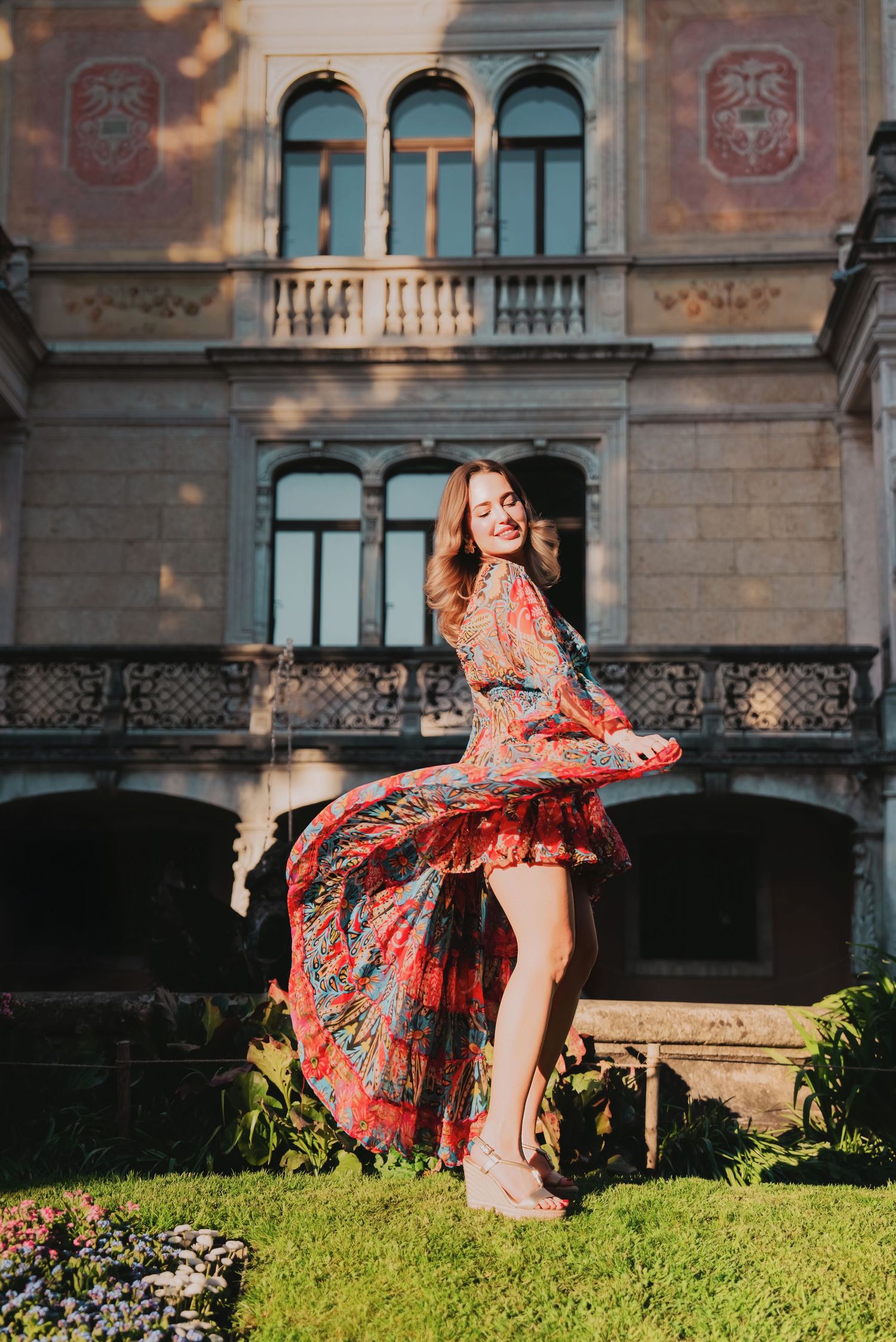 Woman in flowing red floral dress twirling in front of ornate historic building on sunny day.