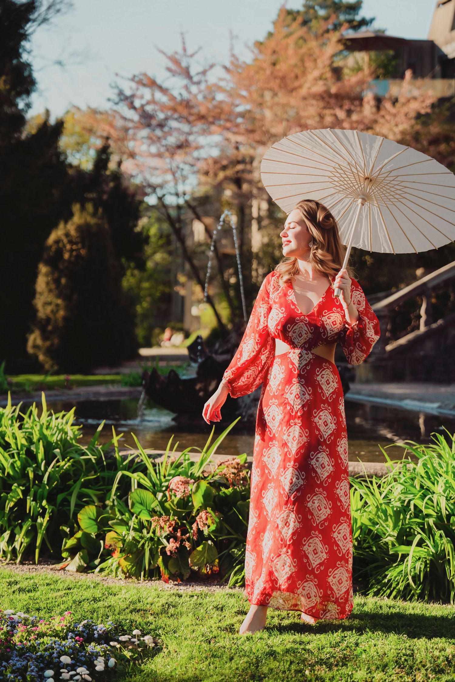 Woman in red floral maxi dress holding white parasol in lush garden by fountain.