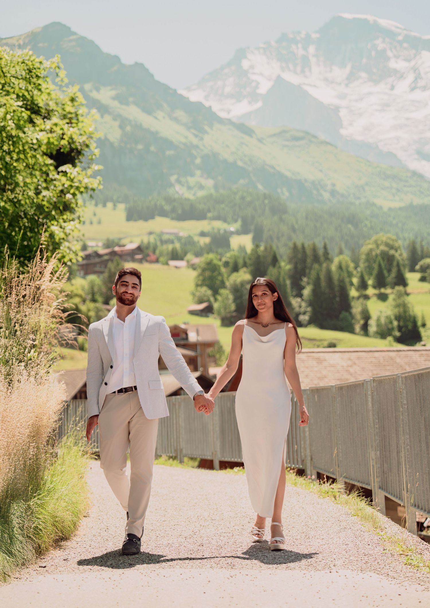 Elegant couple holding hands walking on a mountain path with stunning Swiss Alps scenery behind them.