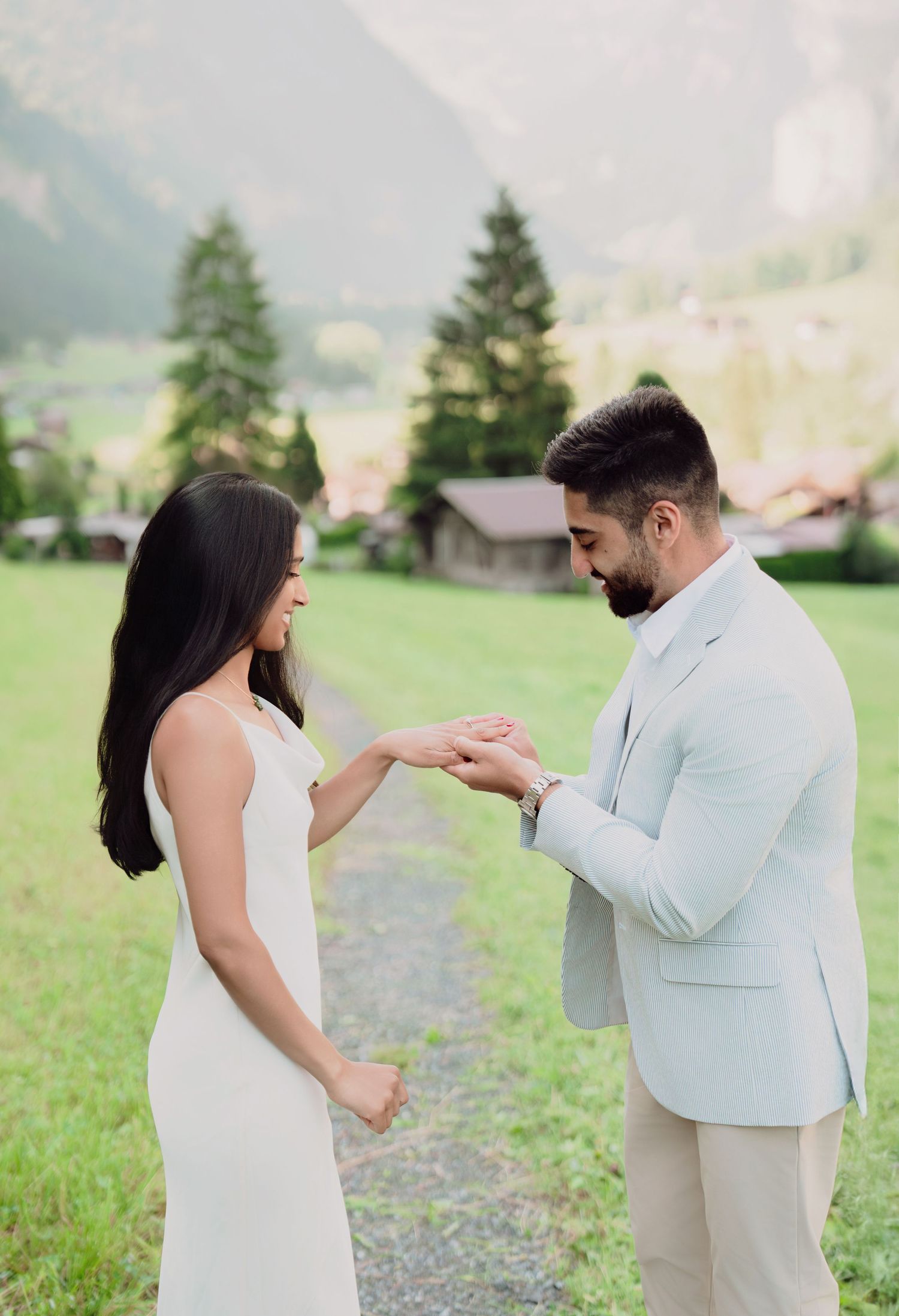 Man placing engagement ring on woman's finger in Swiss alpine meadow with mountain village backdrop.