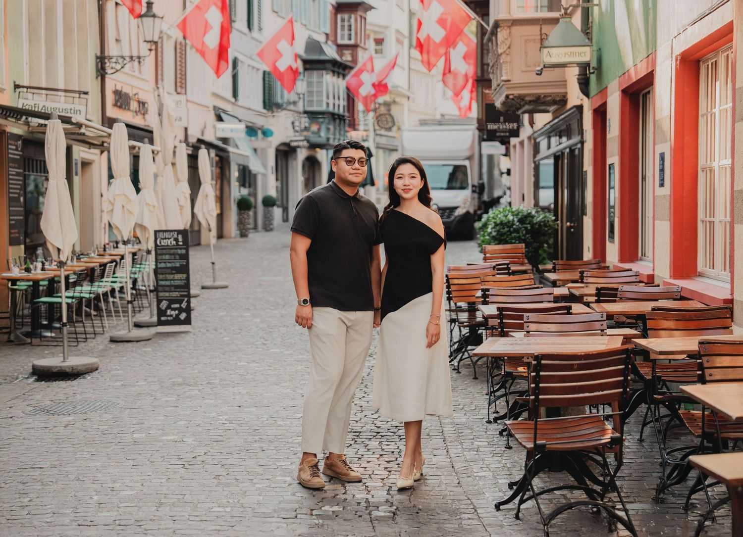 Couple posing together on a charming Swiss cobblestone street with outdoor cafe seating and Swiss flags.
