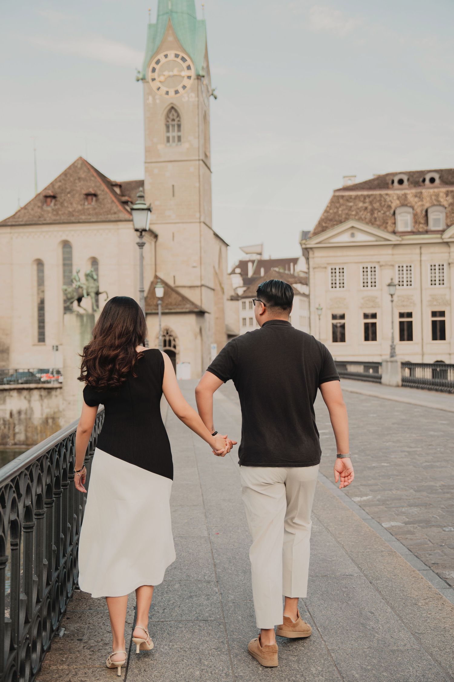 Couple walking hand in hand on a Zurich bridge toward Fraumunster Church on a cloudy day.