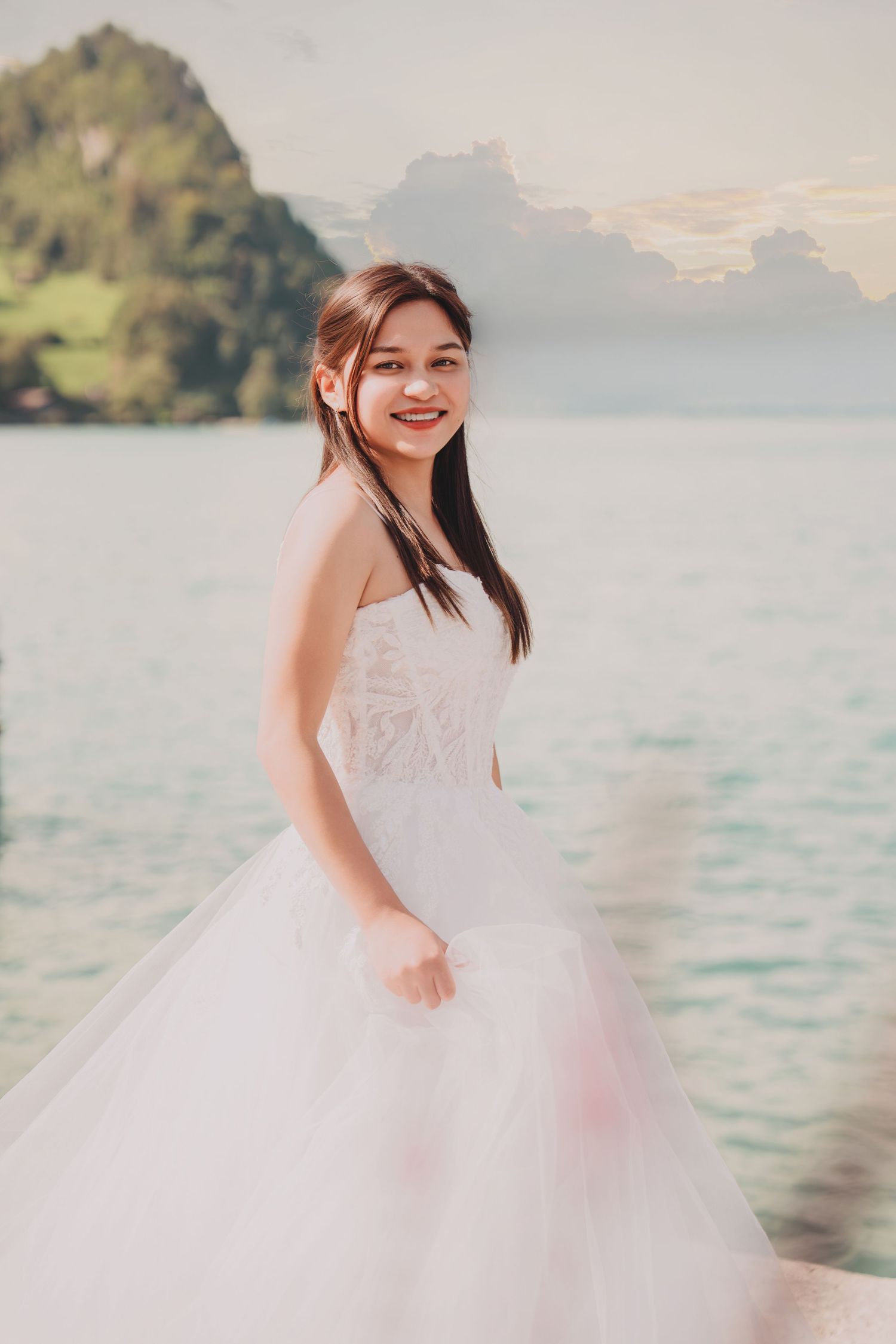 Smiling bride in white strapless lace and tulle gown poses beside a serene alpine lake with mountain views.