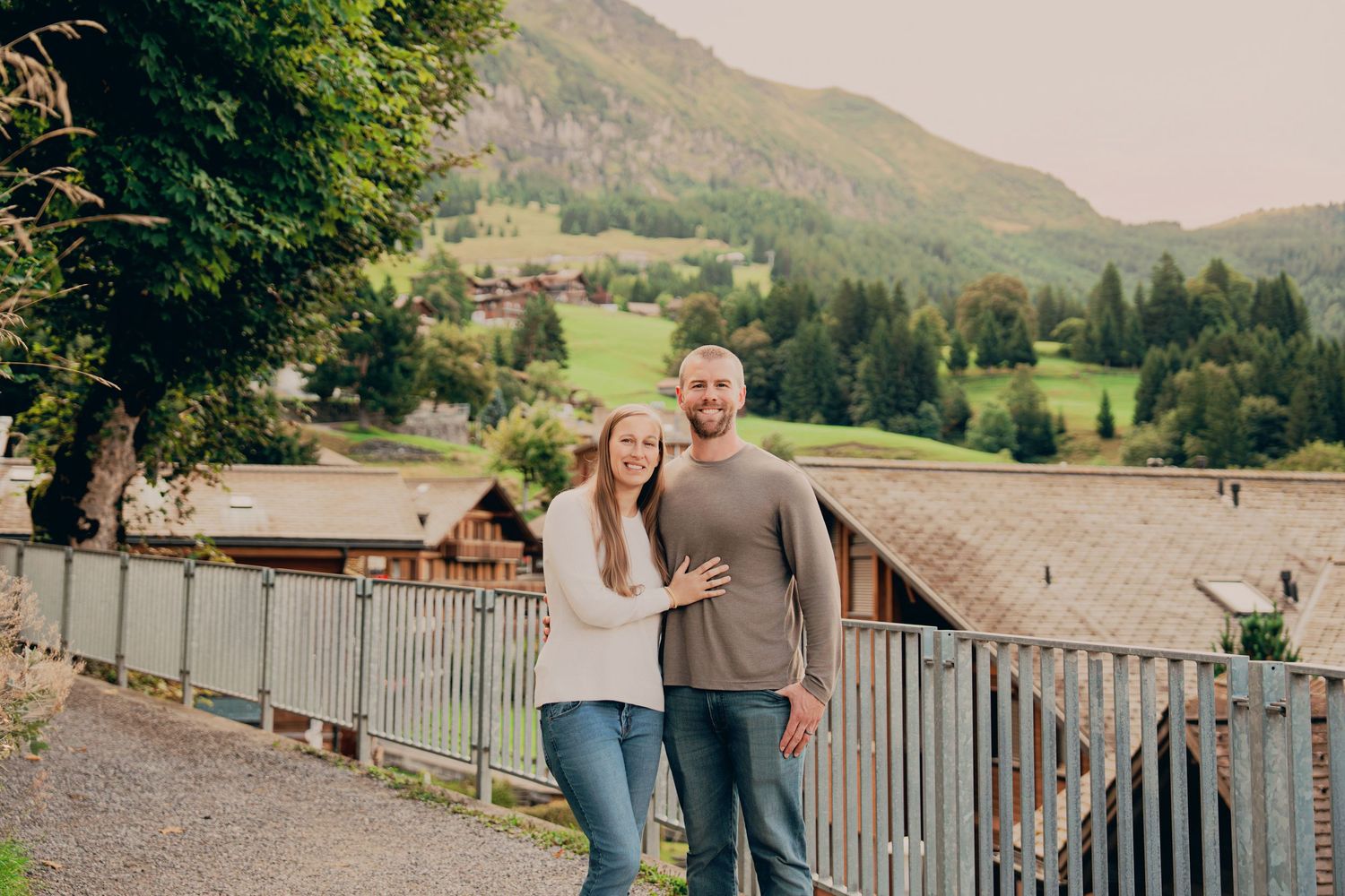 A smiling couple stands together on a scenic alpine path with mountain village views behind them.