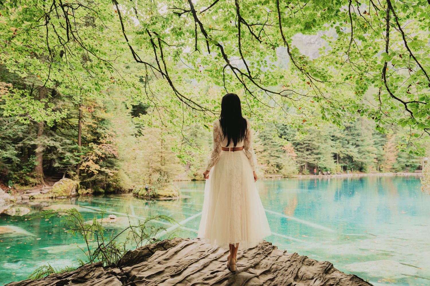 Woman in white lace dress stands on rock overlooking vivid turquoise lake in lush forest.