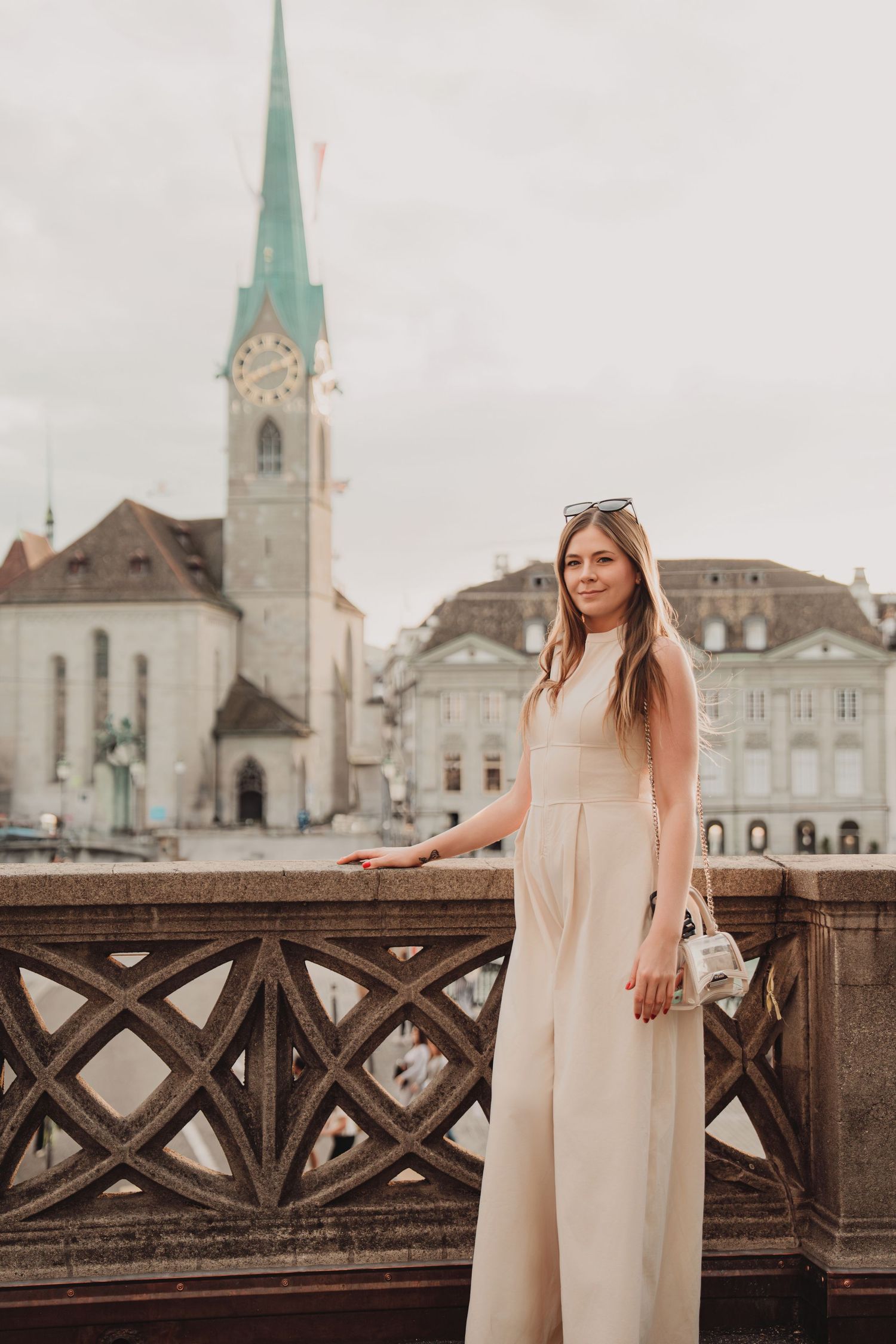 Stylish woman in beige jumpsuit poses on Munsterbrucke bridge with Fraumunster Church tower, Zurich.