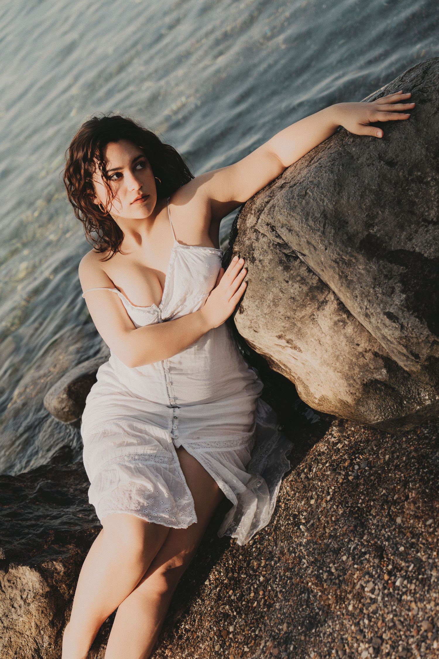 Woman in wet white dress posing against large rocks by the sea.