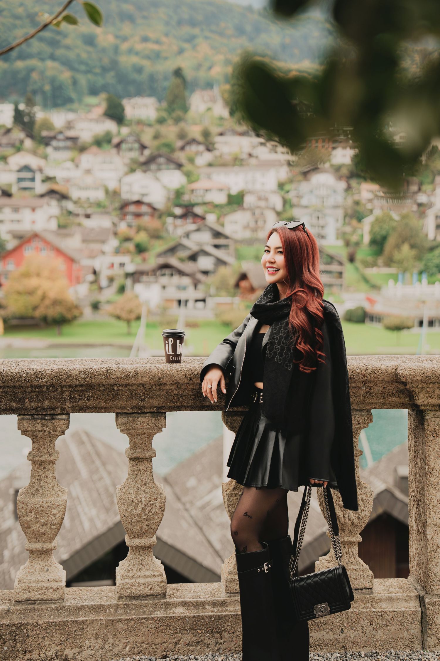 Smiling red-haired woman in black leather outfit holds iced coffee while leaning on stone balustrade with Swiss village view.