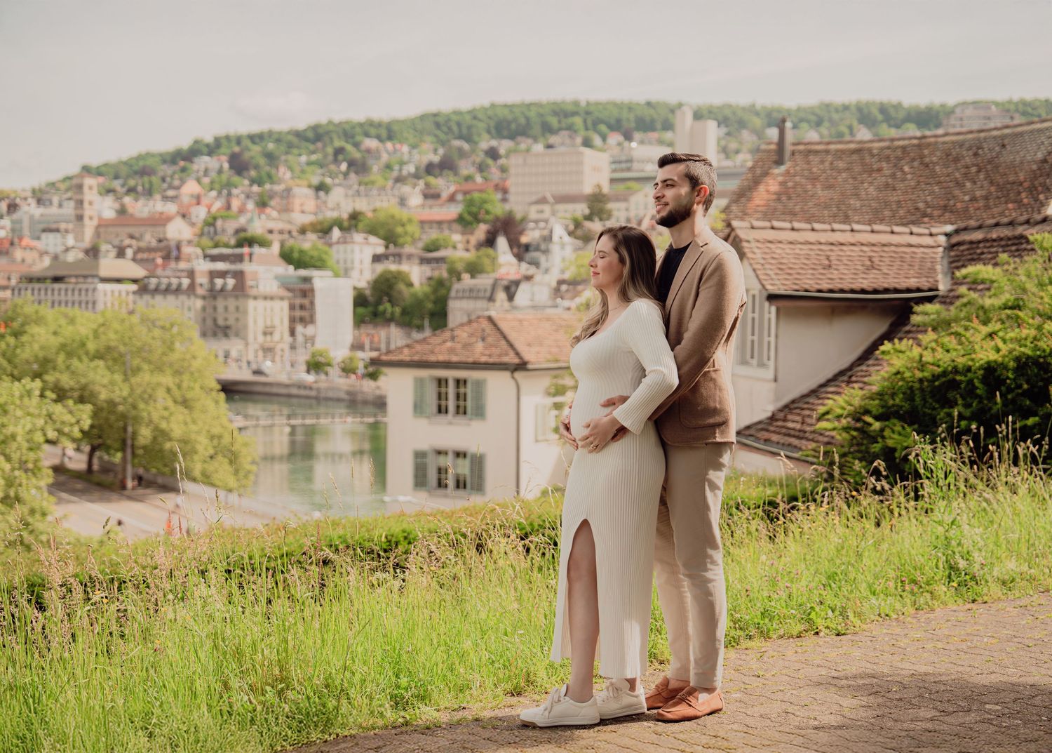 Expecting couple embraces on scenic Zurich hillside overlook with city rooftops and green hills in background.