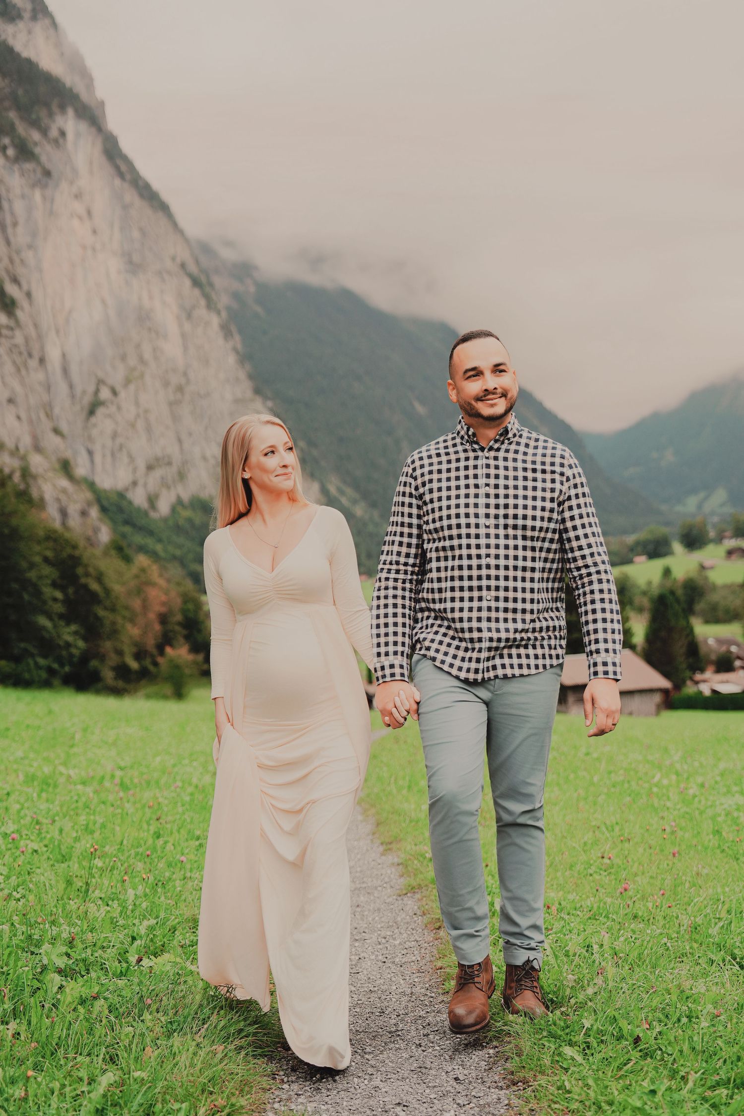 Smiling pregnant couple strolls hand in hand on path through lush green Swiss Alpine valley.