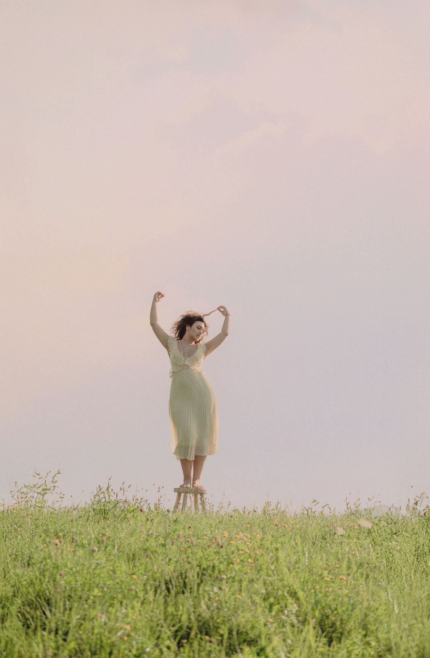 Woman in pale yellow dress standing on stool in flower field with arms raised against pastel sky.