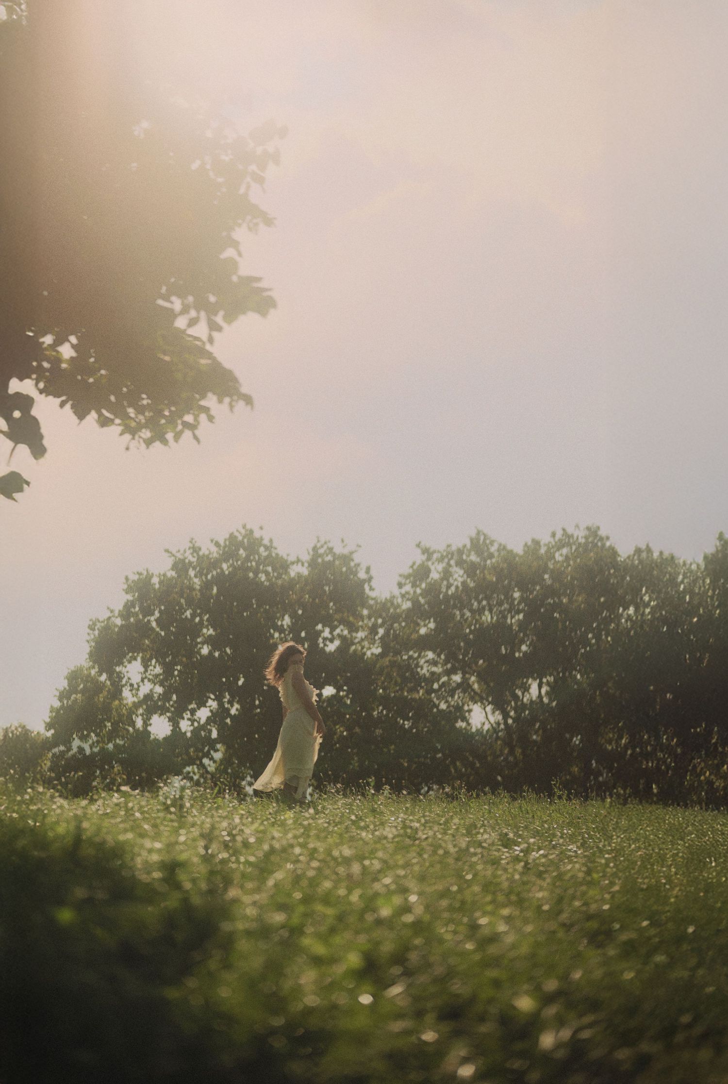 A woman in a white dress stands in a sunlit meadow surrounded by lush green trees.