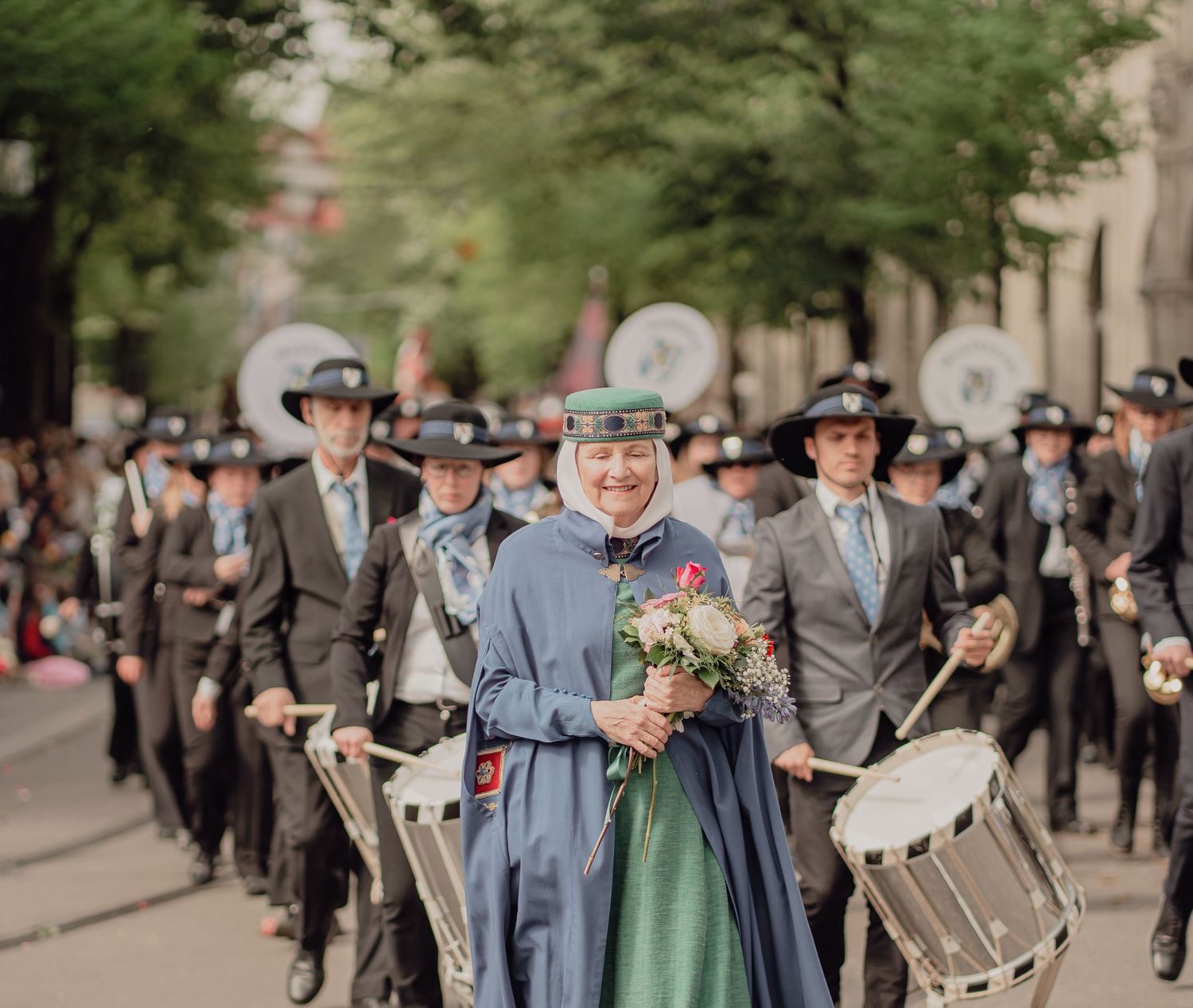 Woman in blue traditional dress leads a marching band parade through a tree-lined street.