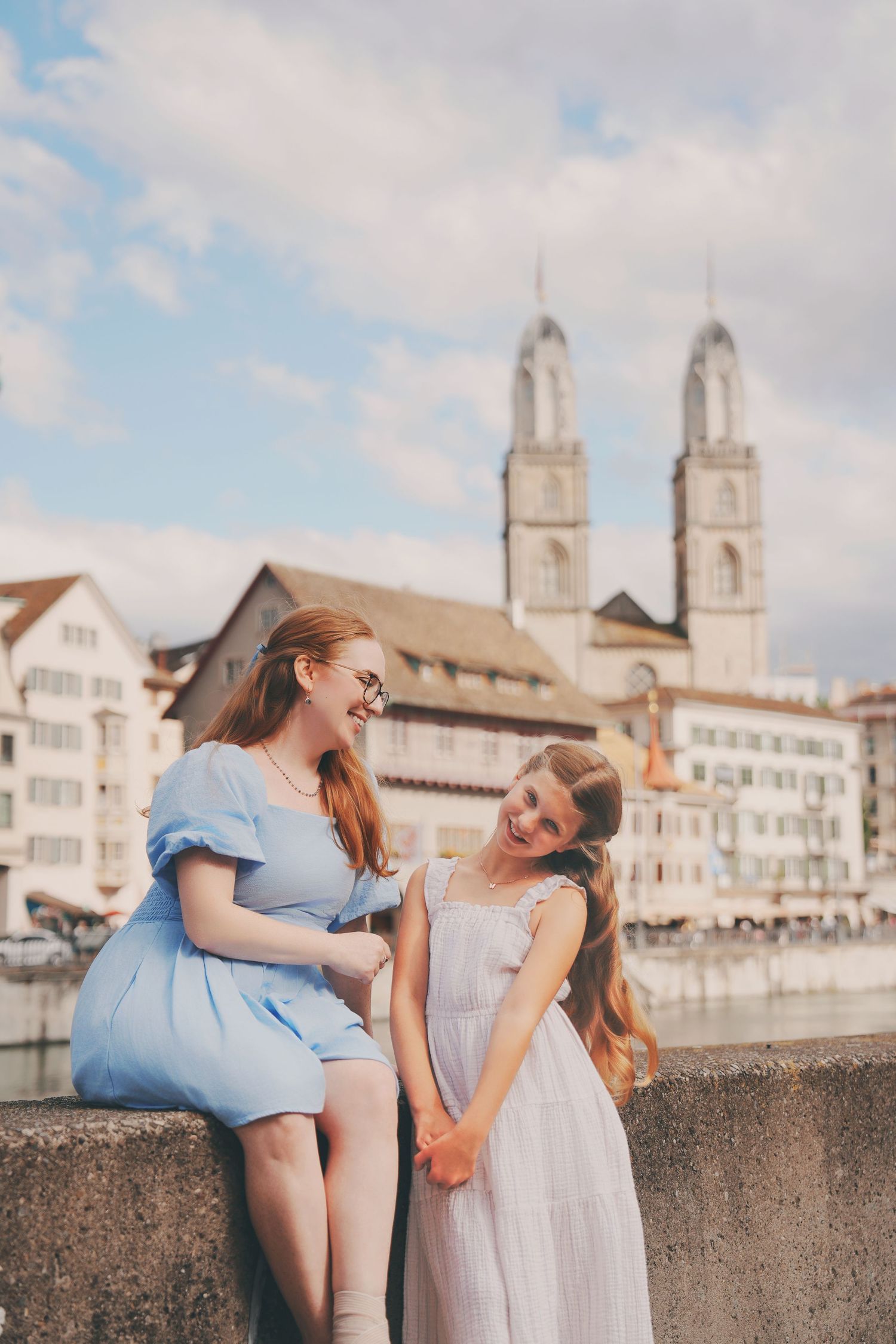Mother and daughter hold hands laughing together on bridge with Zurich skyline behind them.