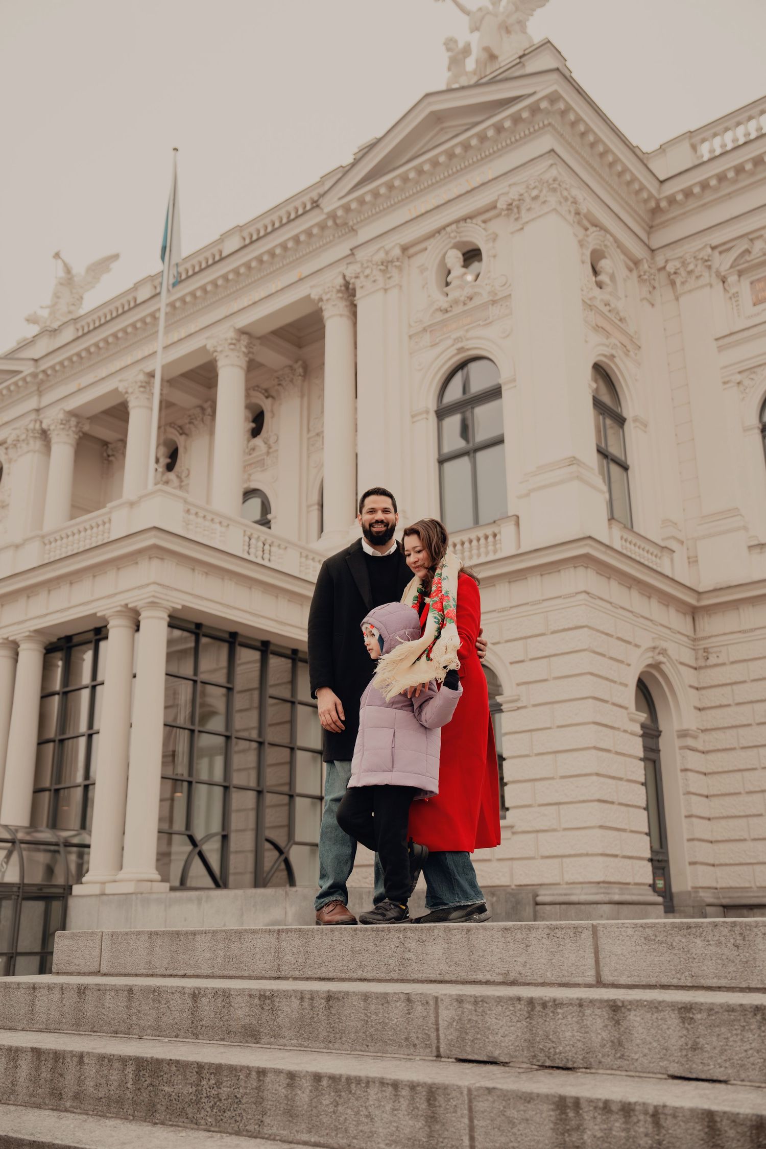 Family of three stands on steps of a grand white neoclassical building, child between parents.