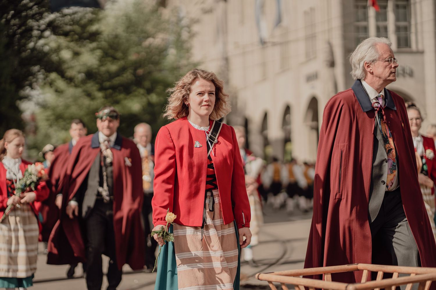 Woman in red jacket and man in red robe march together in a formal outdoor procession.