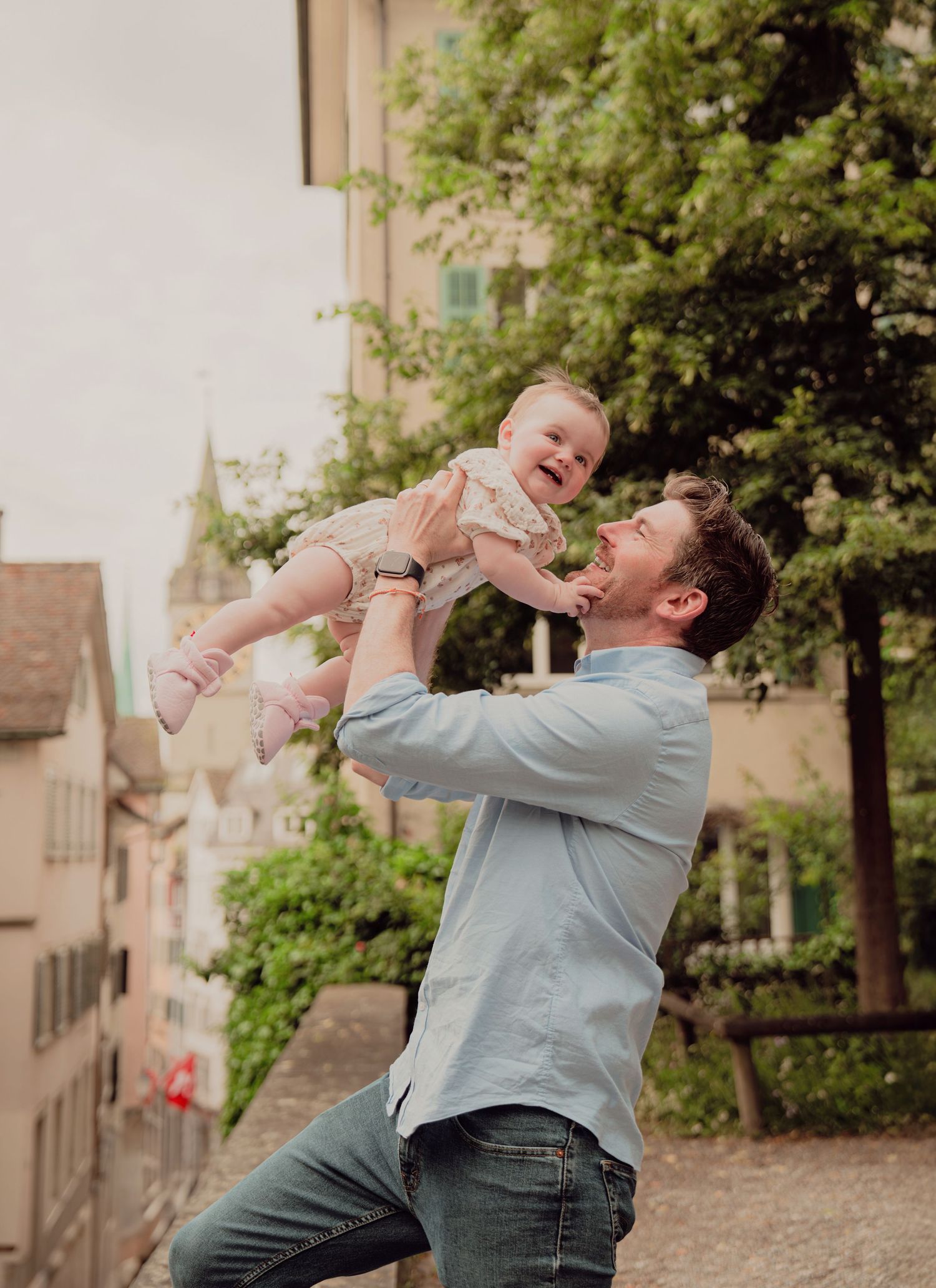 Cheerful father tossing laughing baby in the air in a scenic European town backdrop.