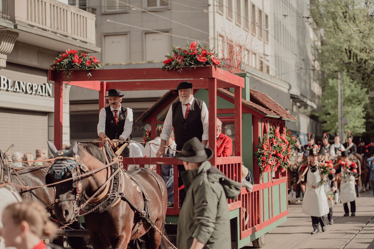 Horse-drawn vintage red tram with men in period costumes rides through a crowded city street parade.