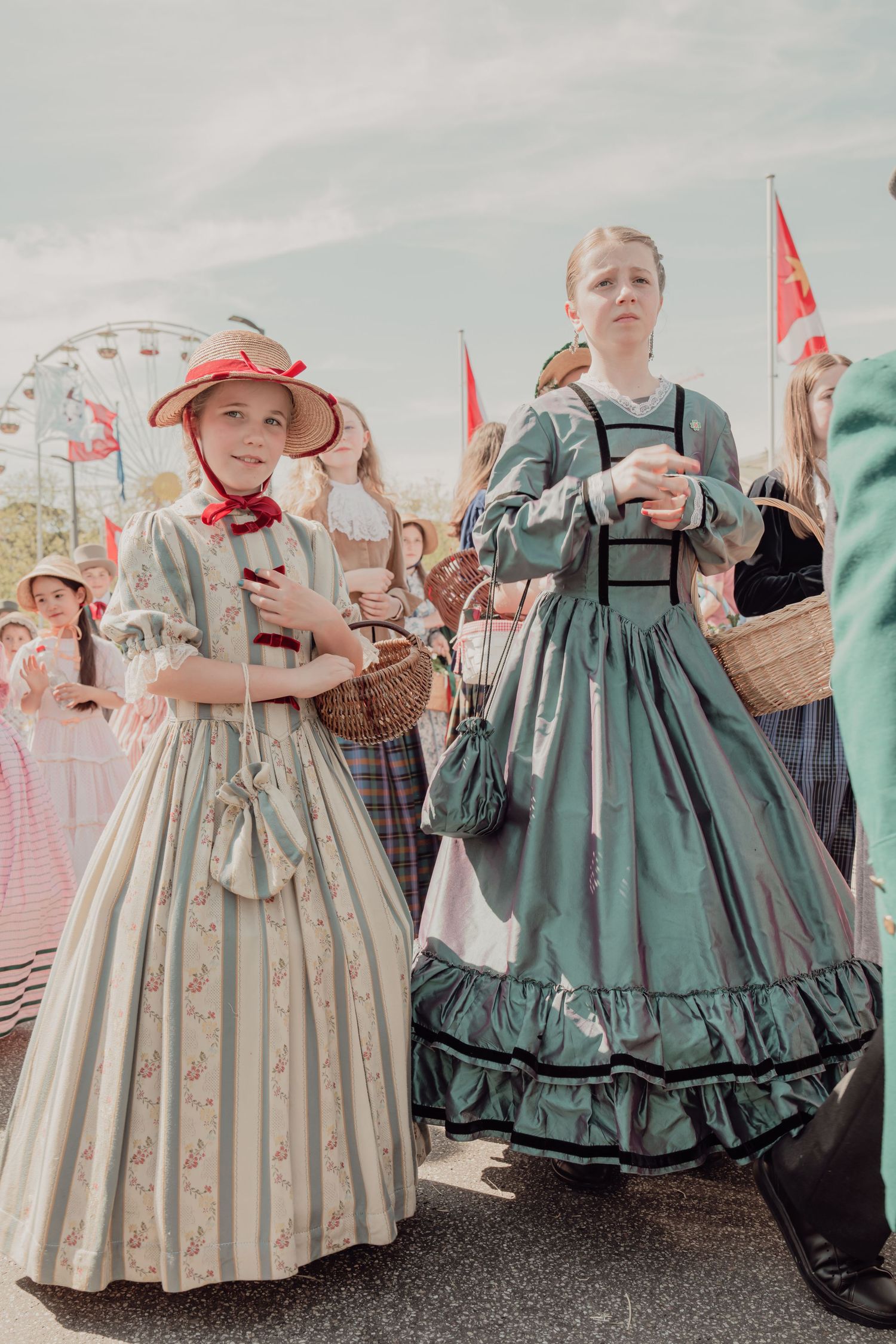 Two girls in Victorian-era dresses with straw hats carry baskets at a traditional Swiss festival parade.