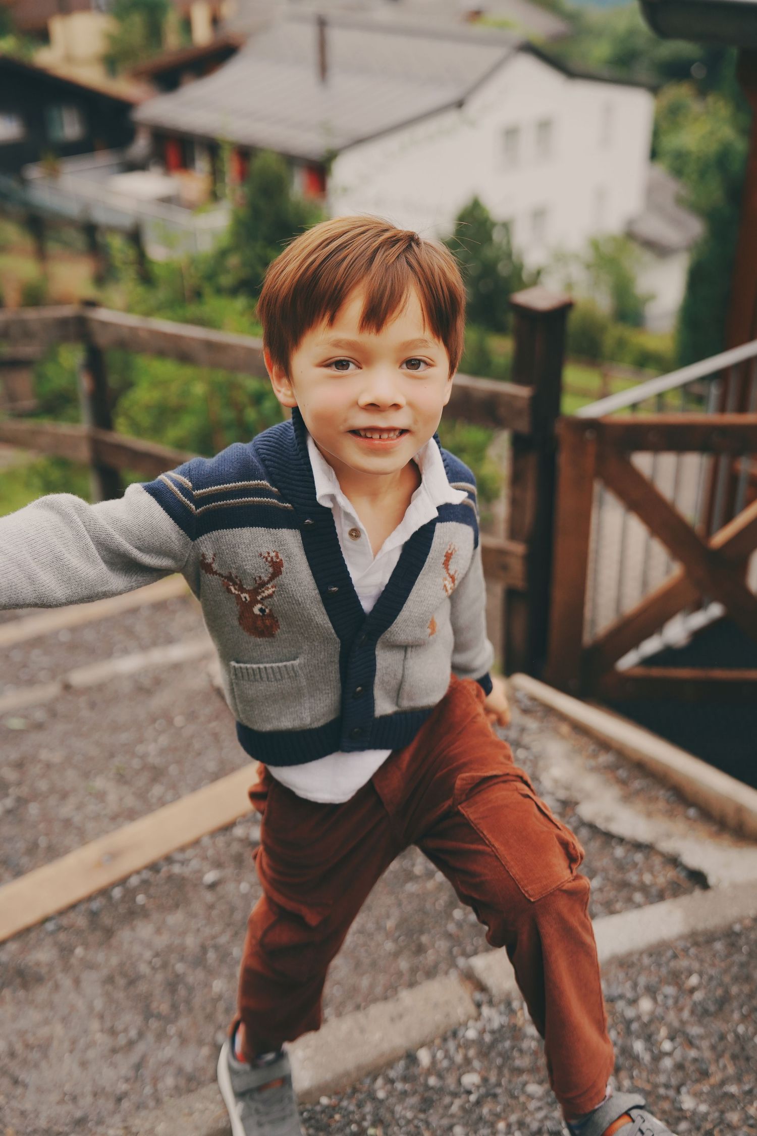 Happy red-haired boy in reindeer cardigan stretches arms wide on outdoor wooden stairs in alpine village.