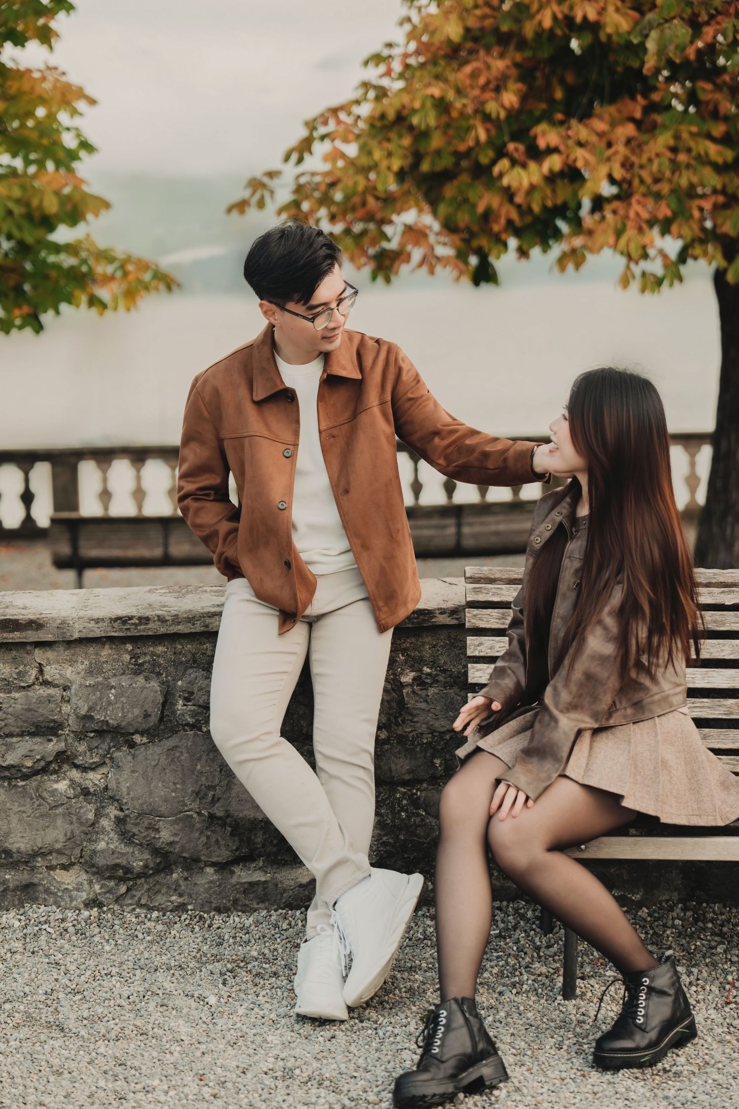 Young Asian couple sharing a tender moment on a bench surrounded by vibrant autumn foliage near a lake.