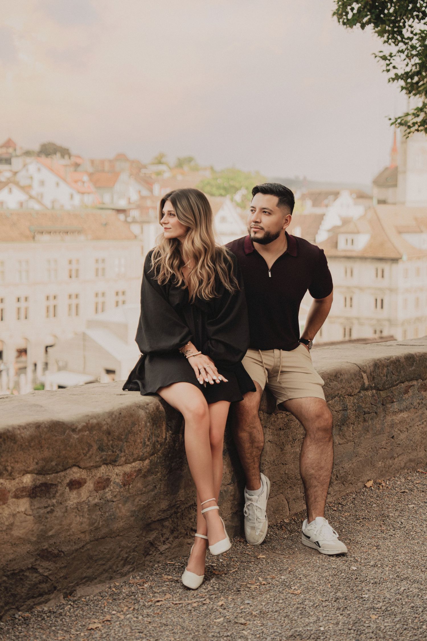 Romantic couple sitting on a stone wall with a panoramic view of Zurich's old town rooftops in the background.