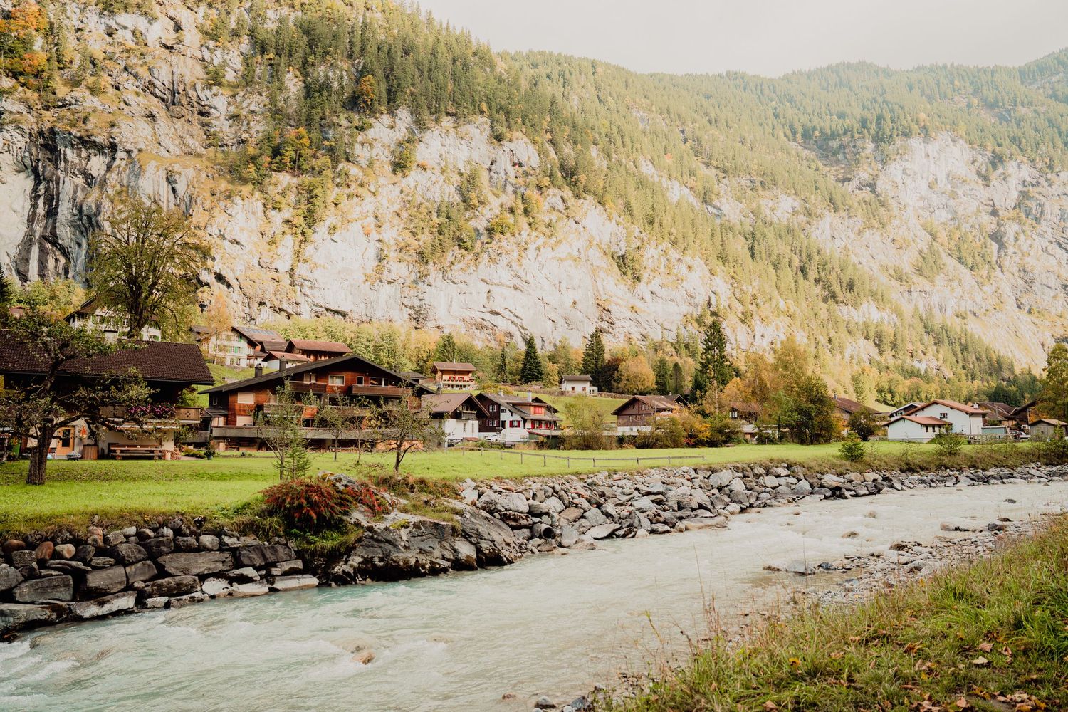 Traditional Swiss wooden chalets nestled beside a turquoise glacial river with dramatic rocky mountain cliffs in autumn.
