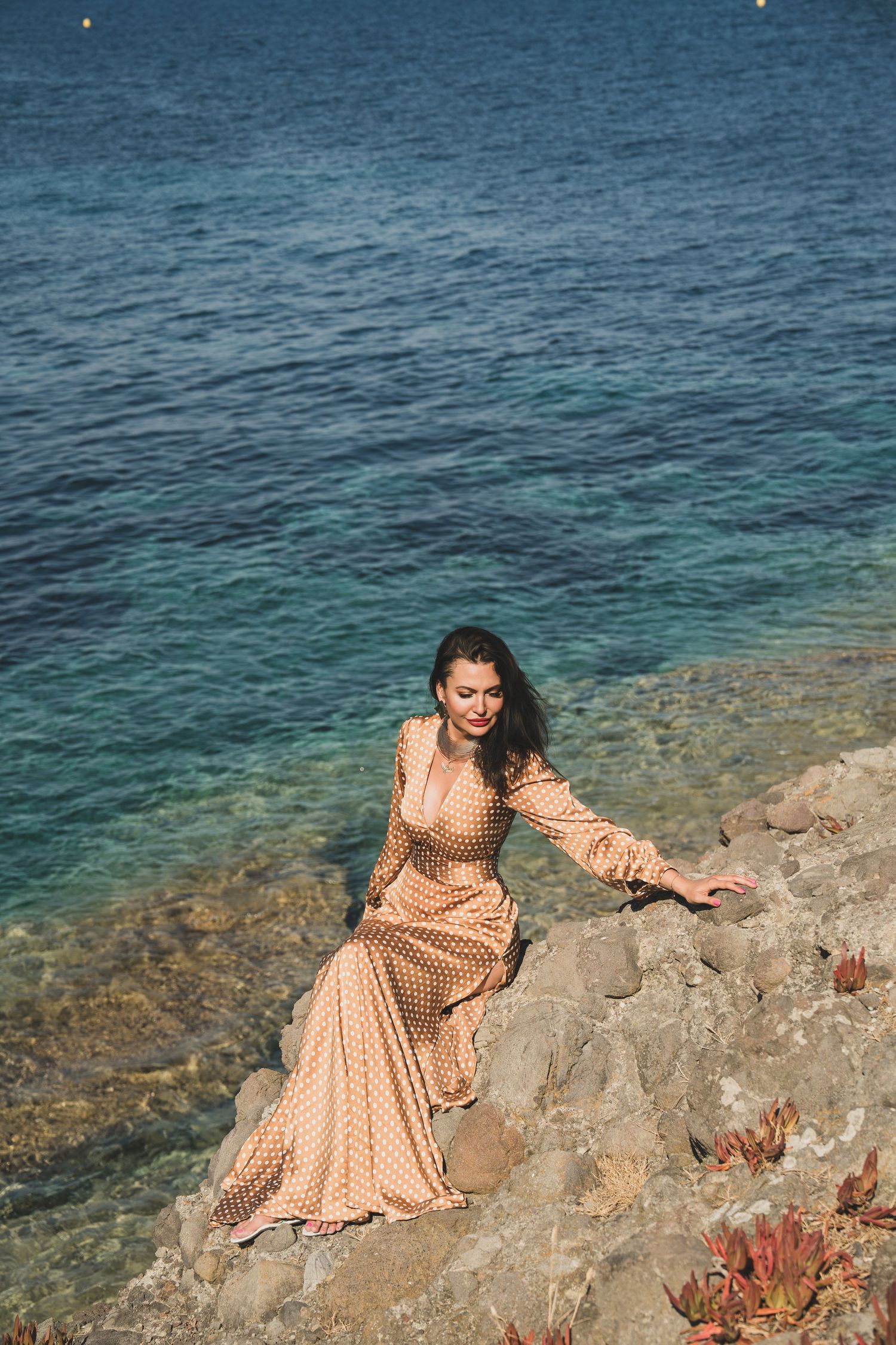 Elegant woman in polka dot dress sitting gracefully on sun-drenched Mediterranean rocks.