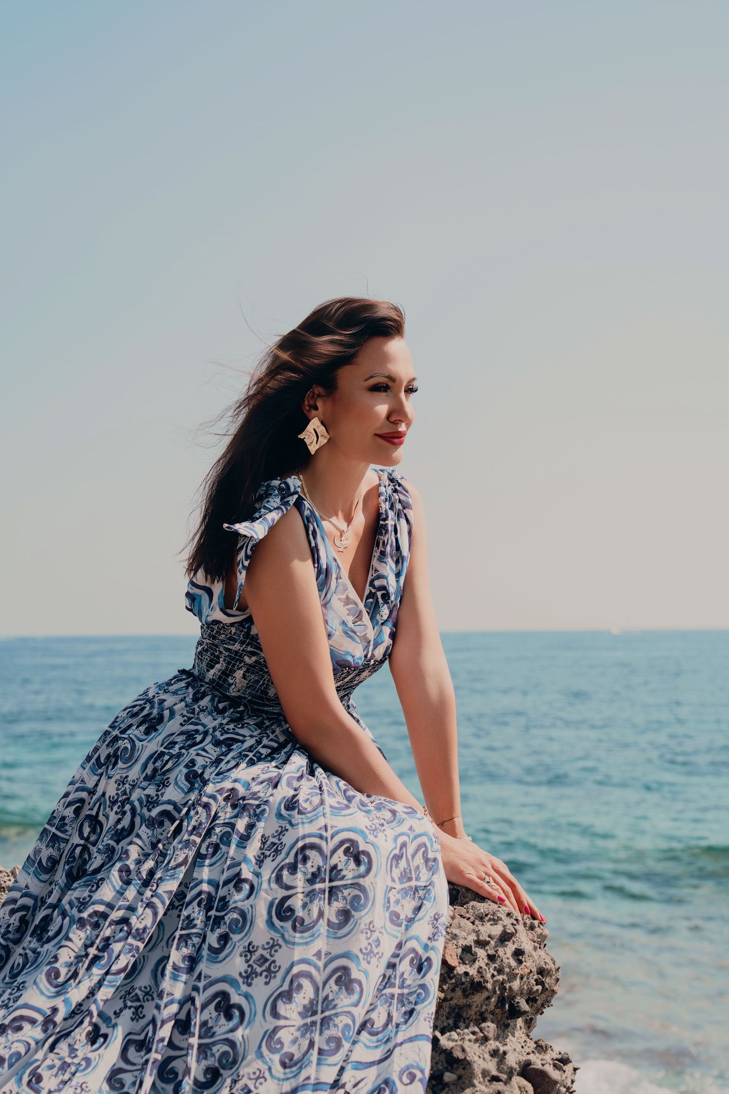 Elegant brunette woman in blue floral print dress sitting on coastal rocks by turquoise sea.