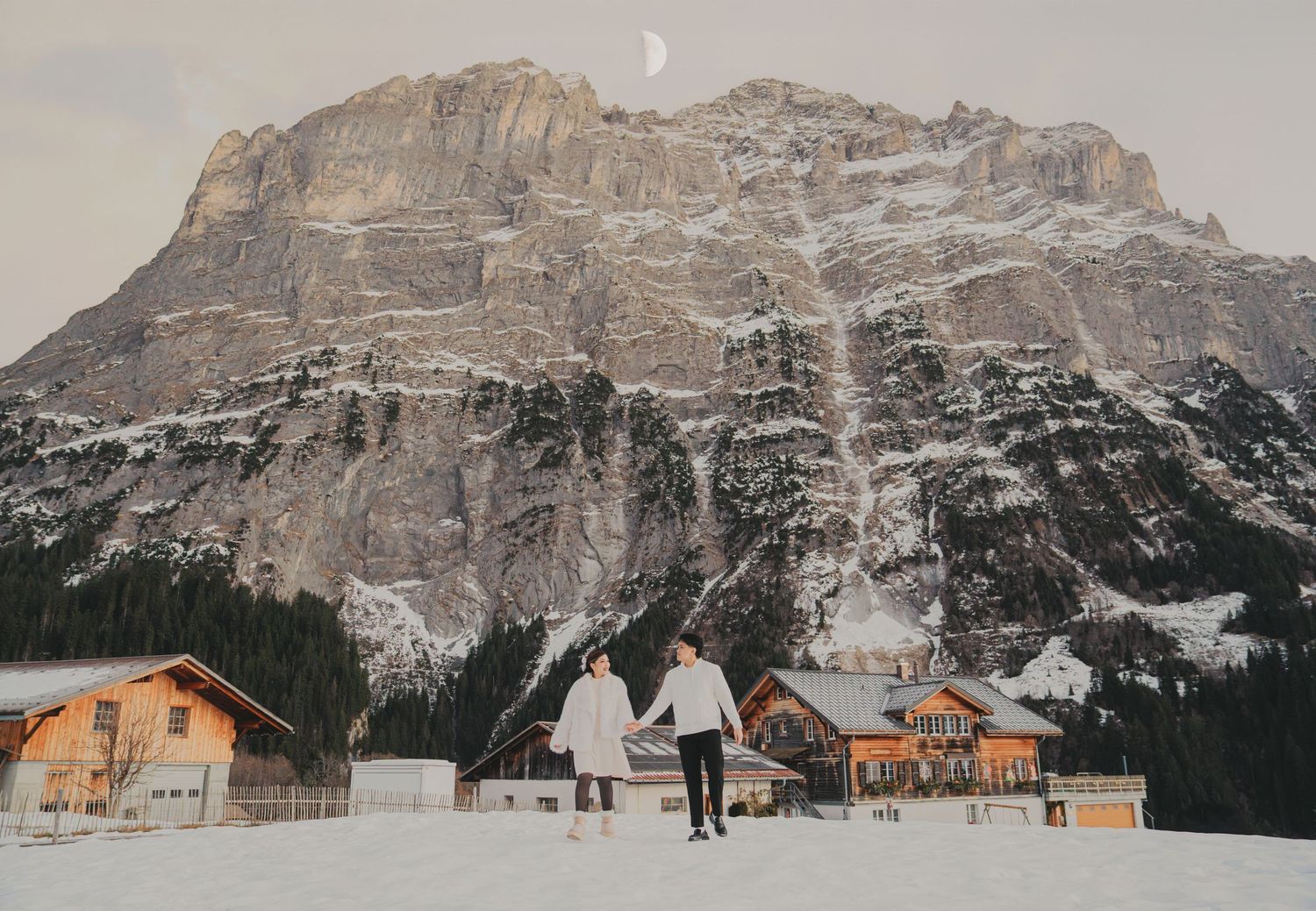 Couple walking hand-in-hand toward snowy Alpine village with dramatic rocky mountain and crescent moon.