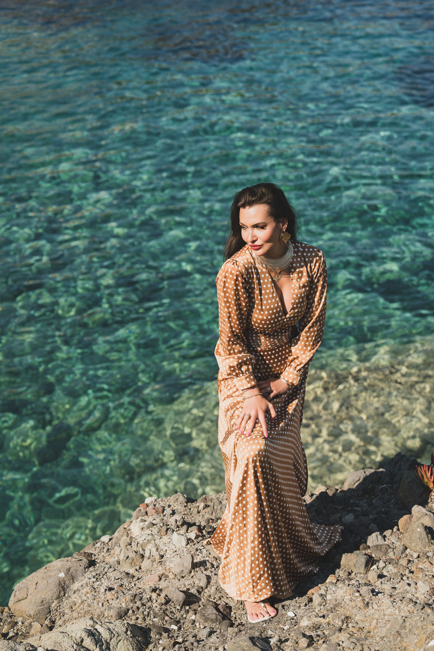 Elegant woman in a gold polka dot maxi dress posing on rocks by crystal clear turquoise water.