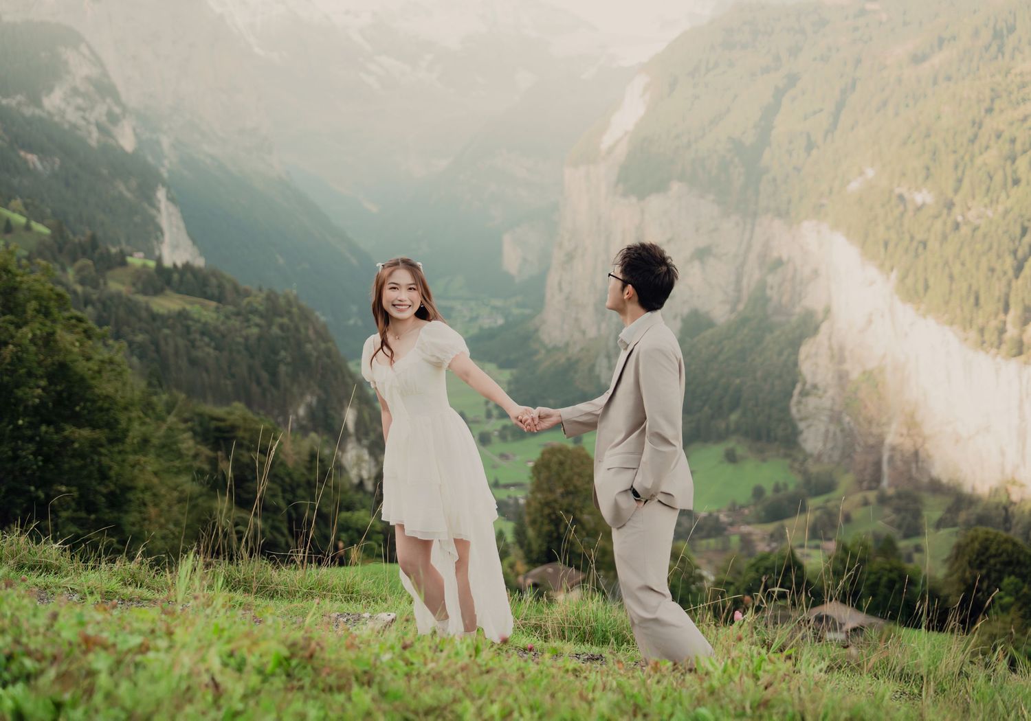 Bride in white dress and groom in beige suit holding hands on Swiss alpine hillside with valley view.