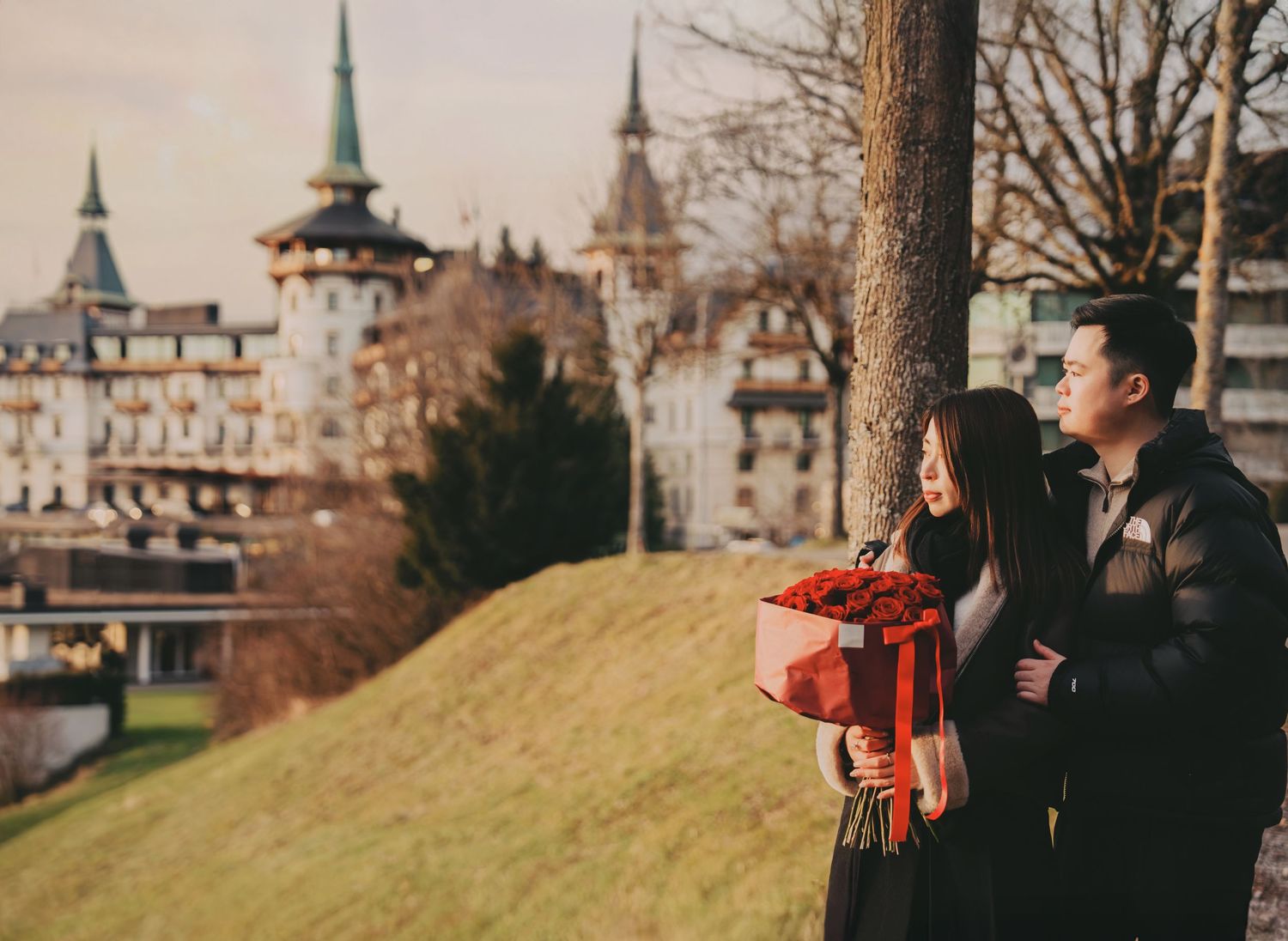 Romantic couple sharing tender moment by tree holding red roses with castle backdrop.