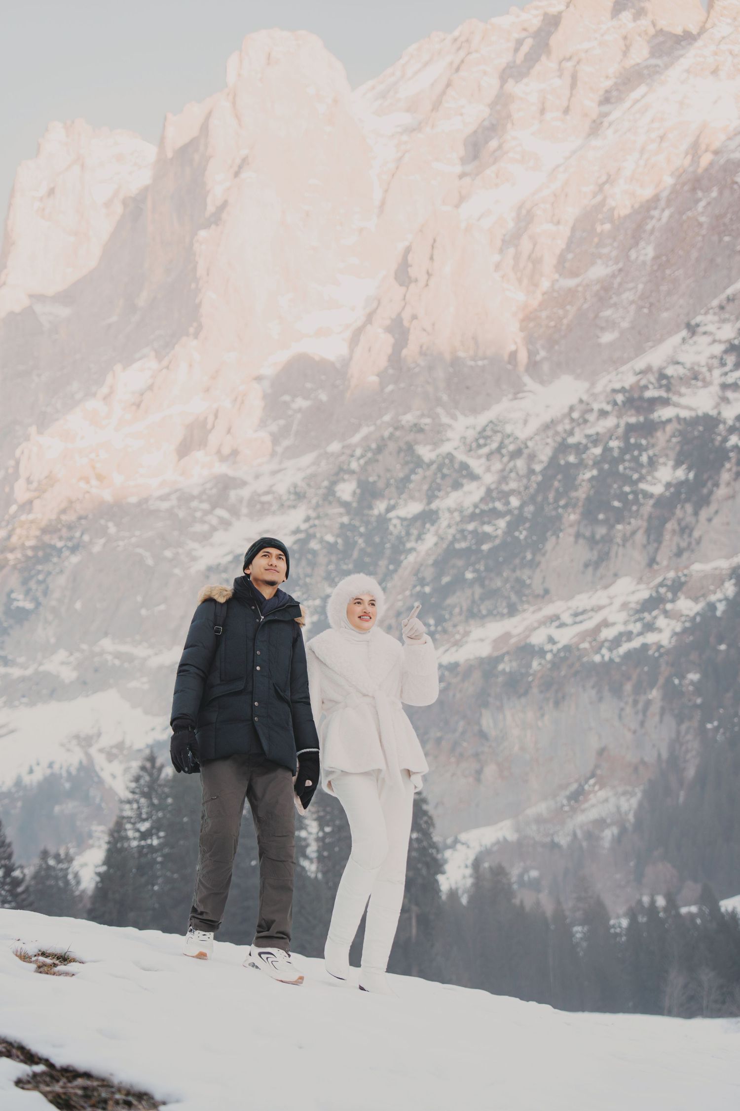 Couple standing together in snowy alpine landscape, woman in white hijab embraced by man in dark coat.