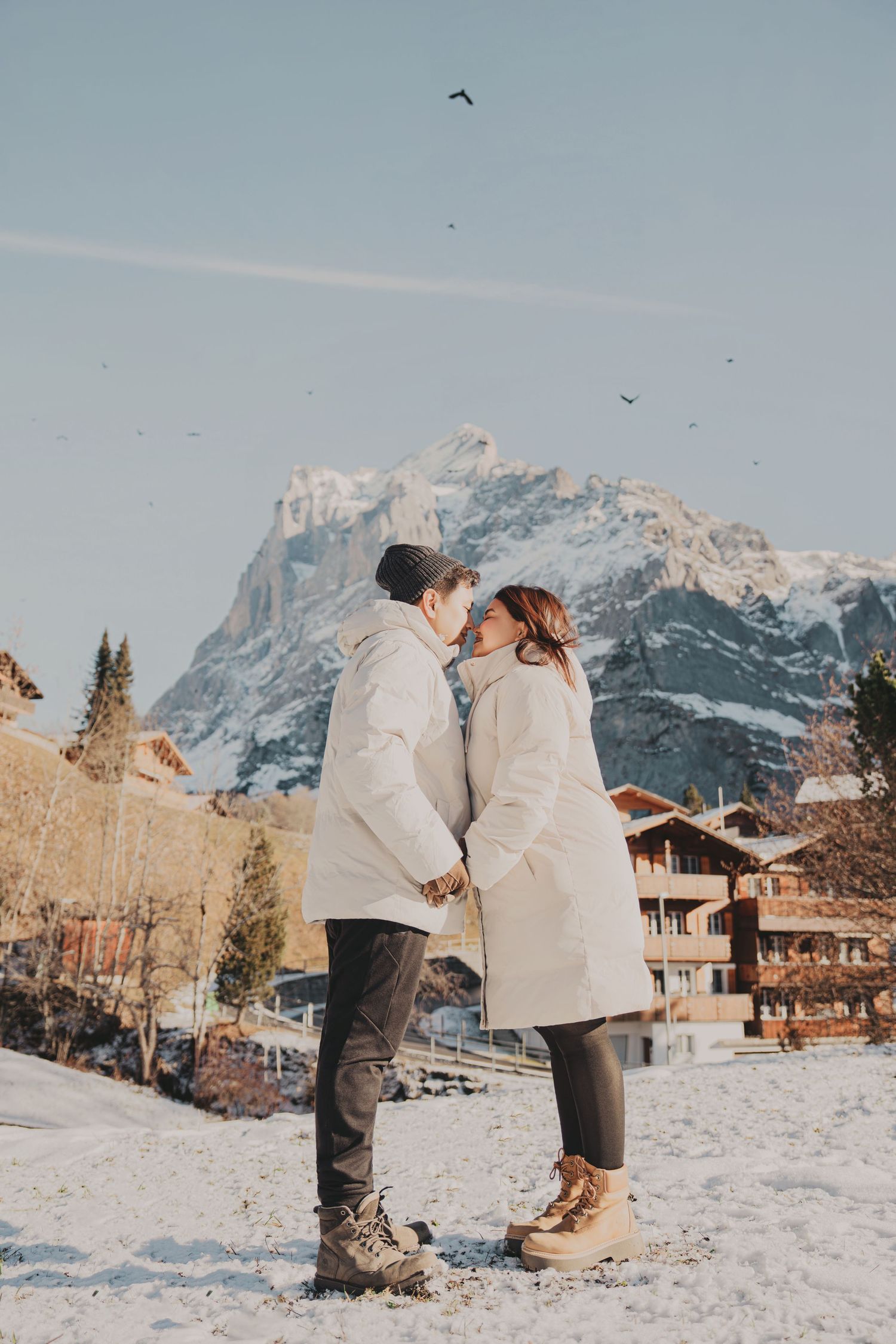 Couple kissing in snowy alpine village with snow-capped mountain backdrop in winter.