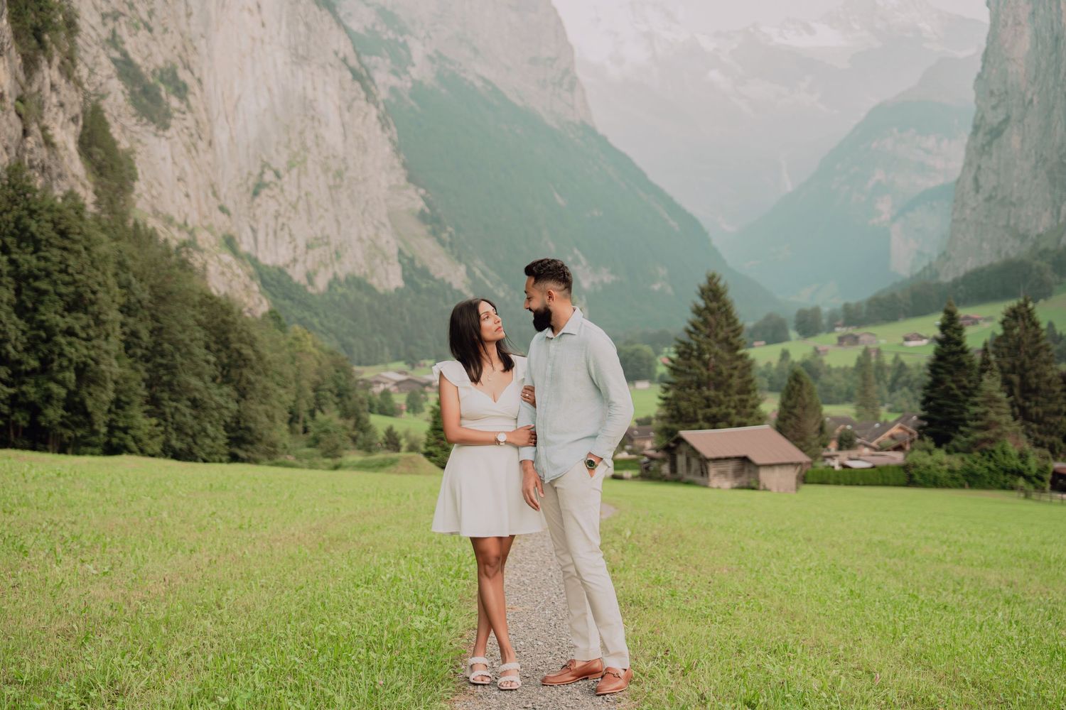 Couple in white dress and light blue shirt holding hands in green alpine meadow with dramatic mountain valley backdrop.