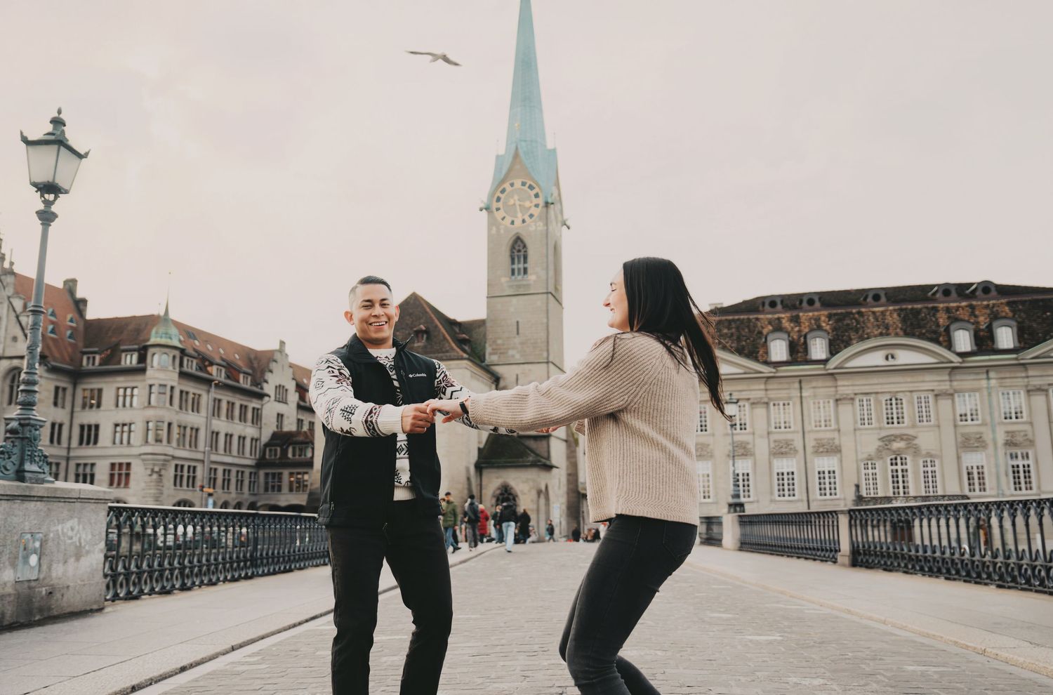 Joyful couple holding hands on a Zurich bridge with historic church spire behind them.