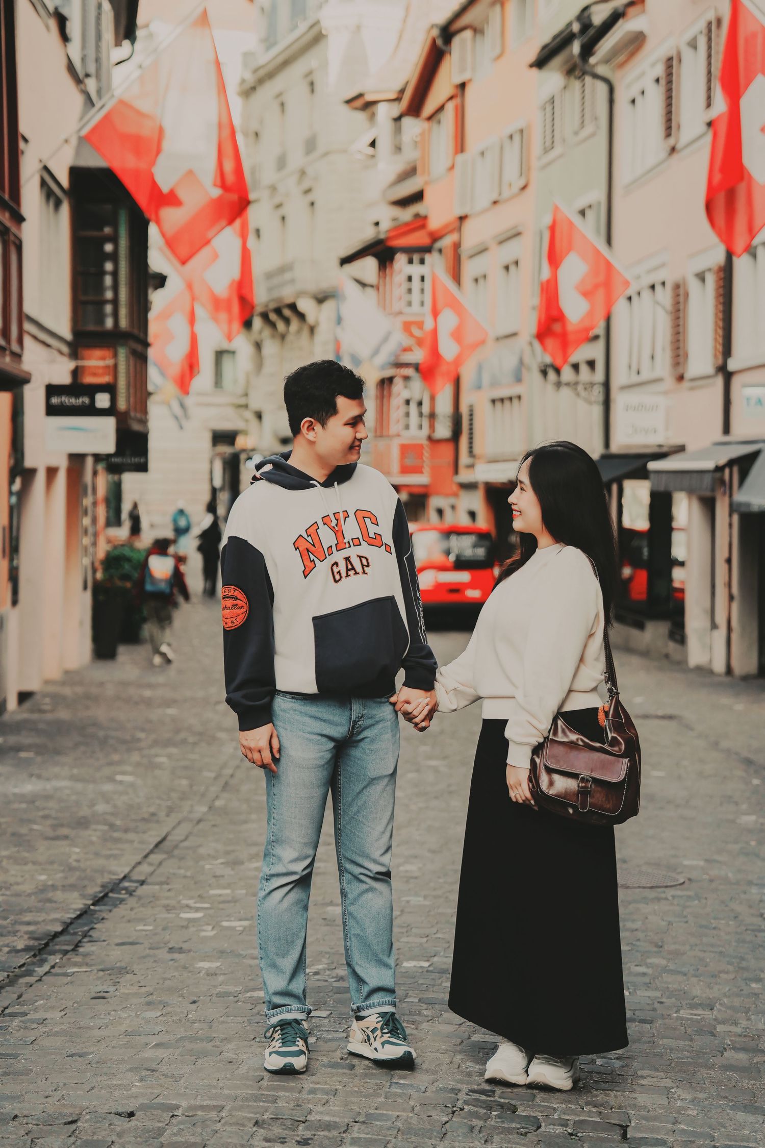 A couple holds hands on a Swiss cobblestone street lined with red and white Swiss flags and historic buildings.