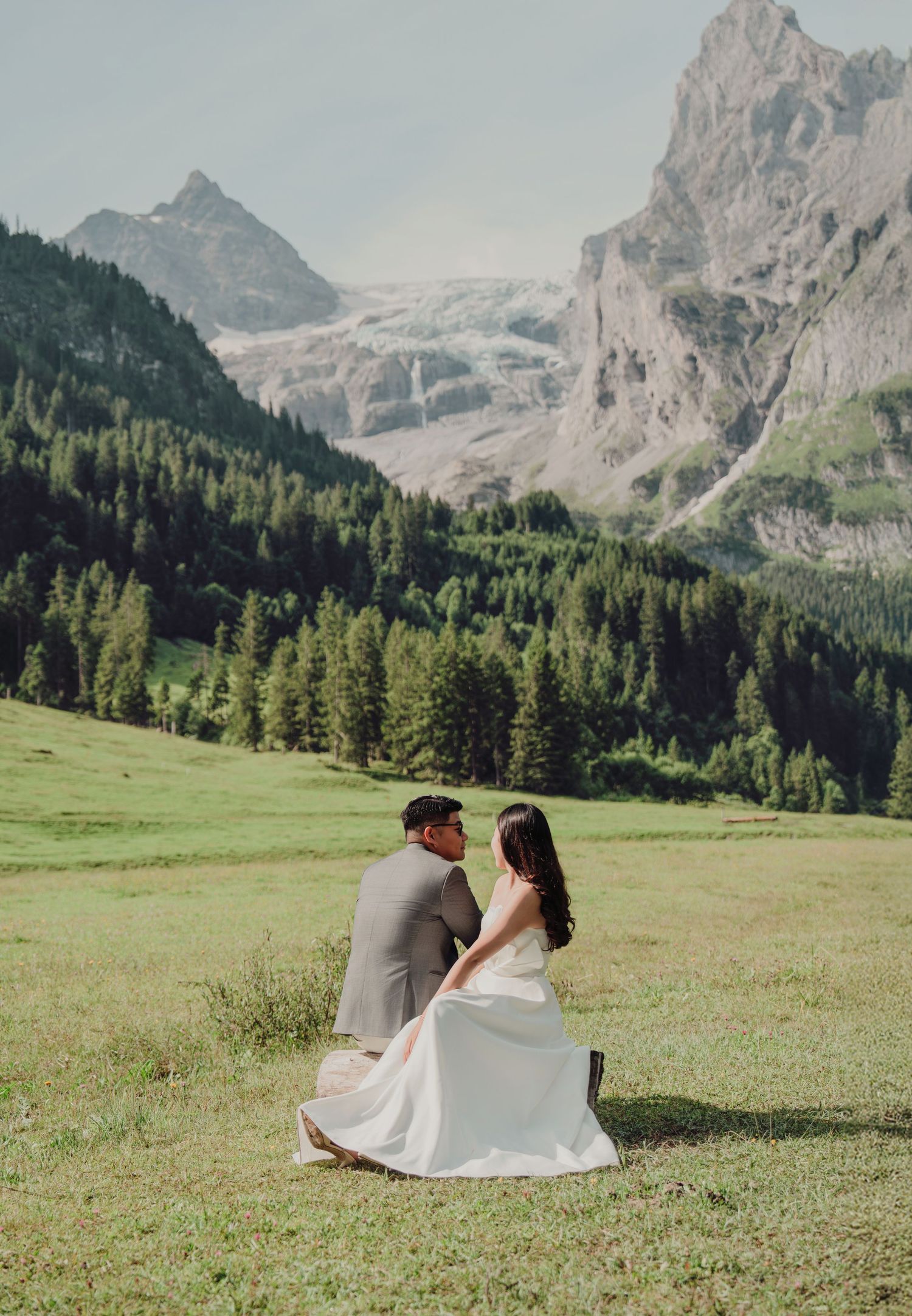 Newlyweds seated on grass in alpine valley with pine forest and rocky mountain peaks behind.