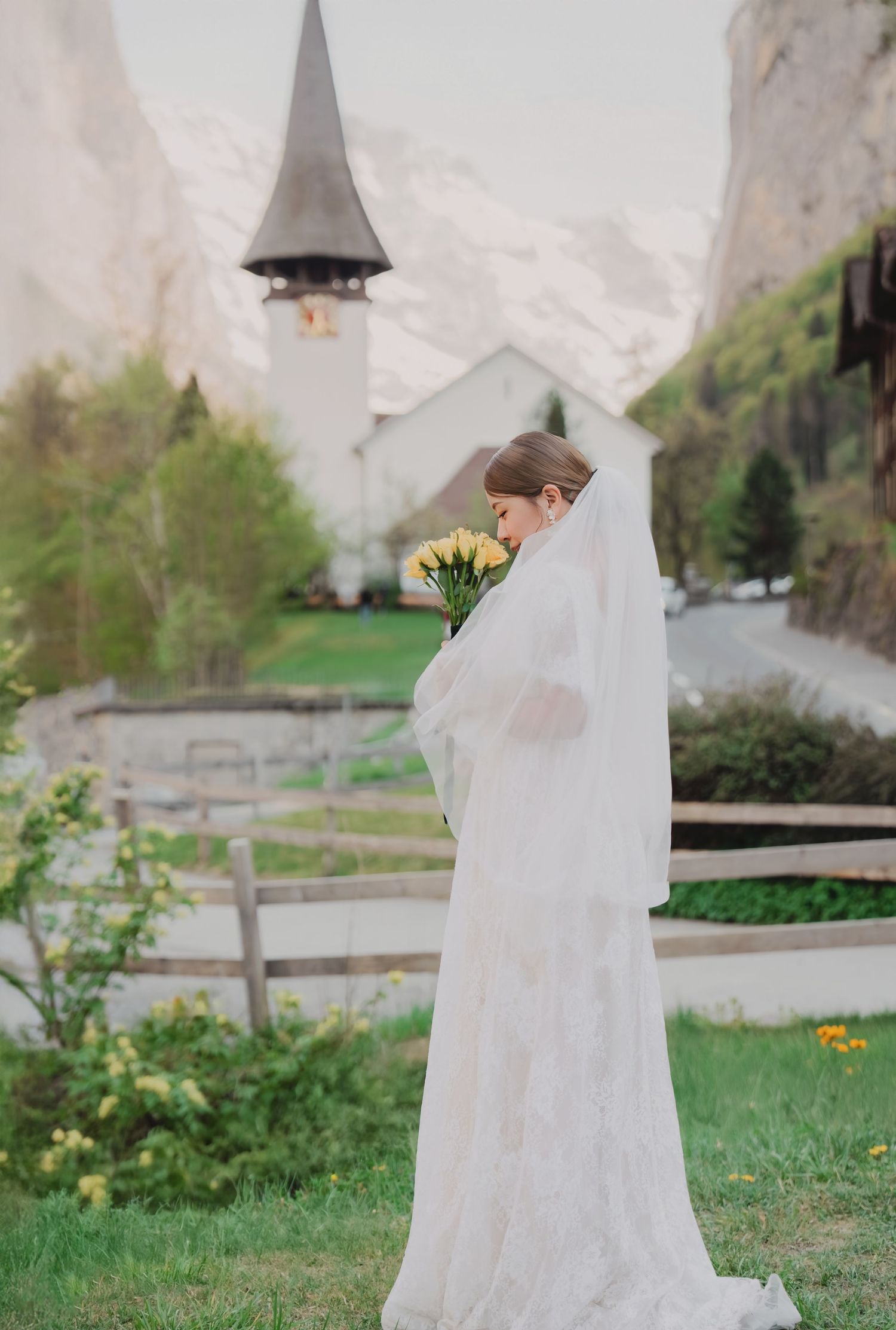 Bride in white lace gown holds yellow tulips before a charming Swiss church steeple.