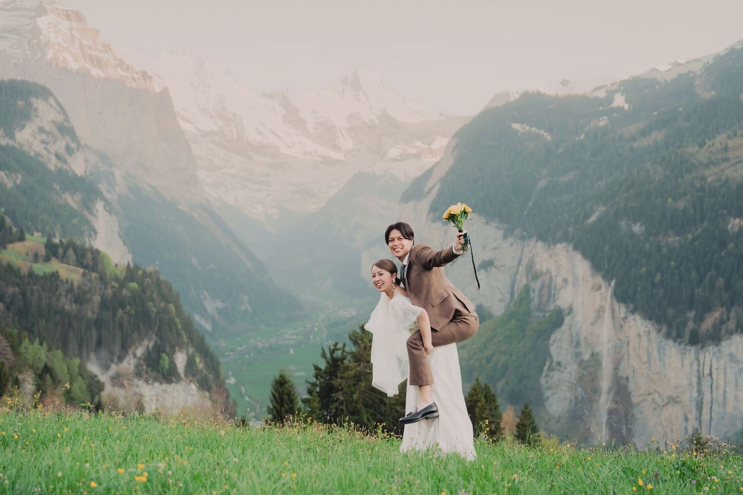 Groom playfully carrying bride on shoulders in alpine meadow with Swiss mountain valley view.