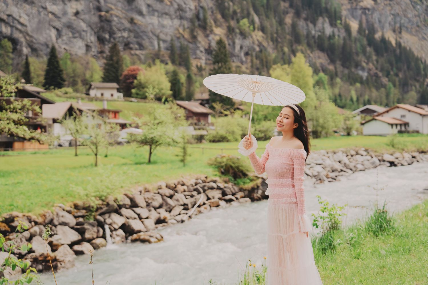 Young woman in pink dress holding white parasol smiling beside a mountain river in Swiss village.