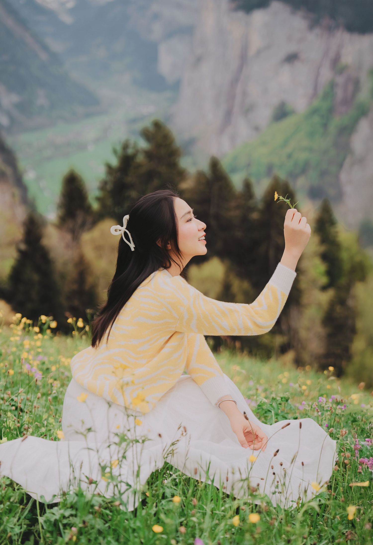 Smiling woman in yellow striped sweater holding wildflower in Swiss alpine meadow with mountain backdrop.