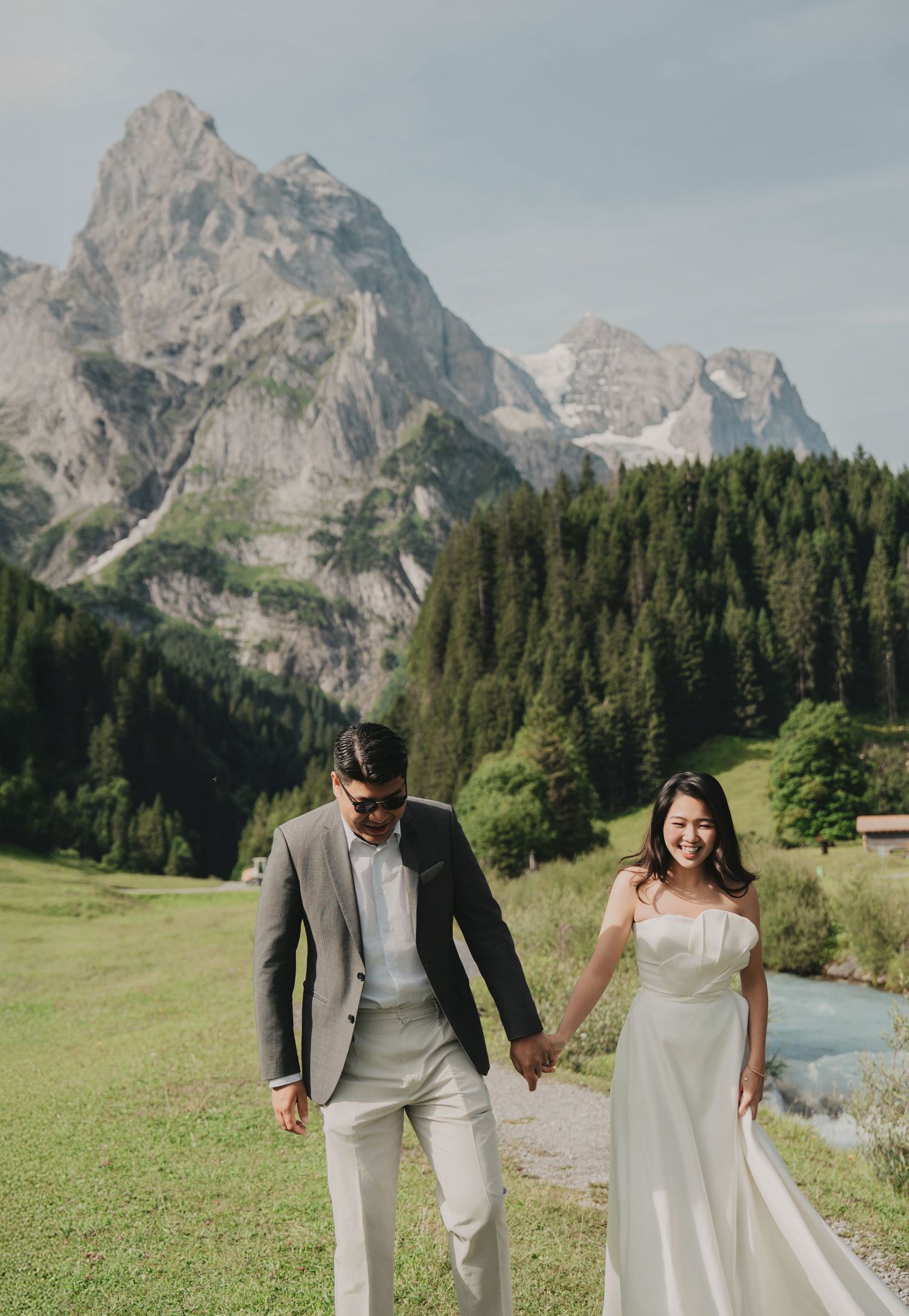 Smiling bride in white gown and groom in gray suit walking together in a stunning alpine valley setting.