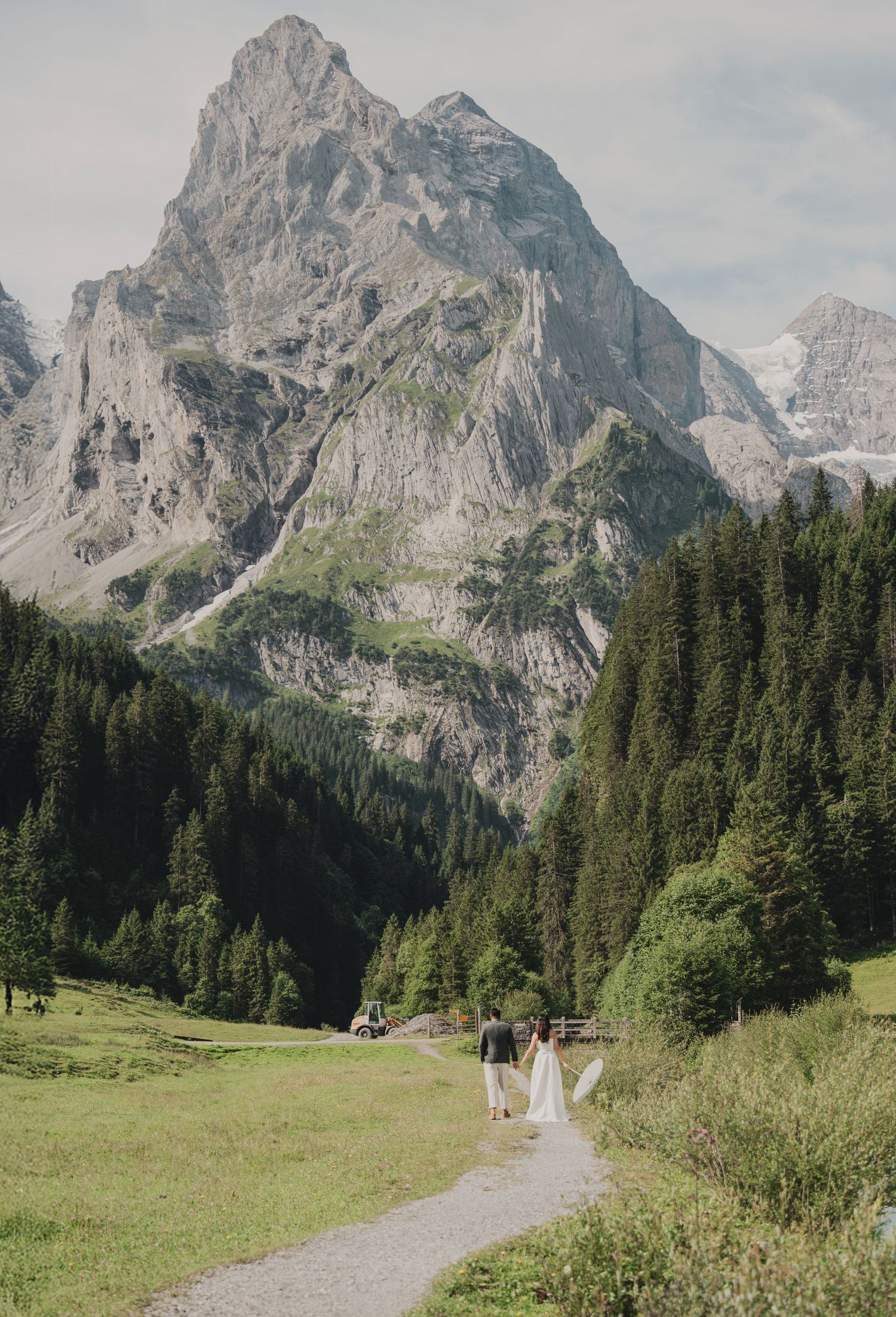 Bride and groom walking a scenic Alpine valley path with towering rocky mountain and evergreen forest backdrop.