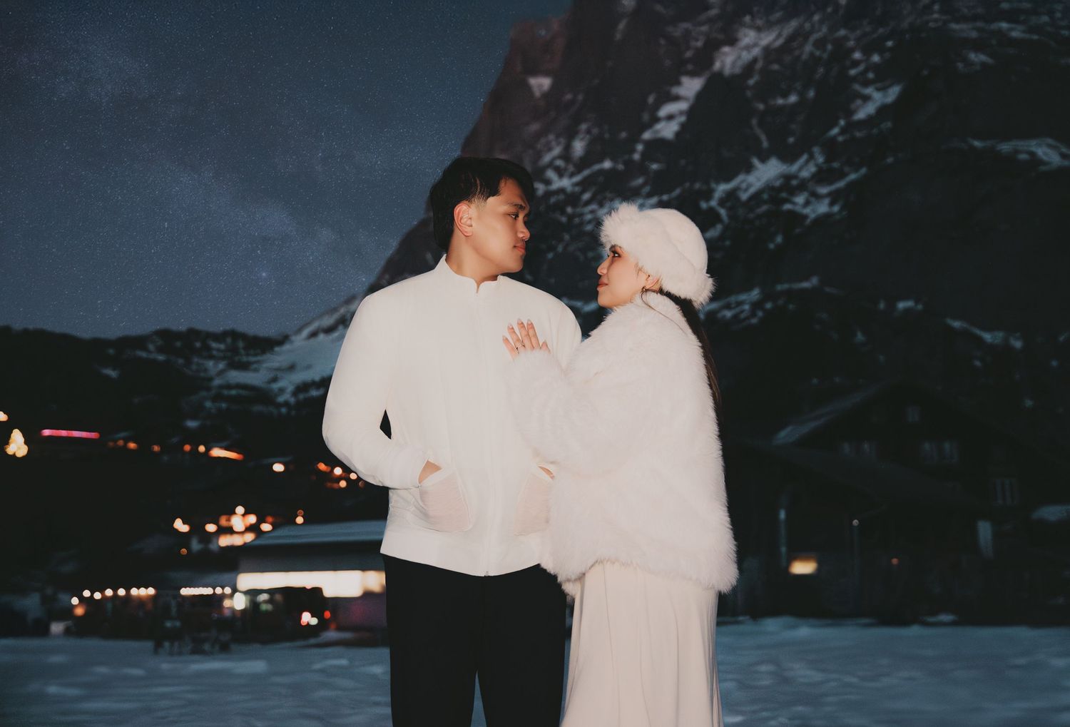 Romantic couple in white winter clothing gazing at each other by snowy mountains.